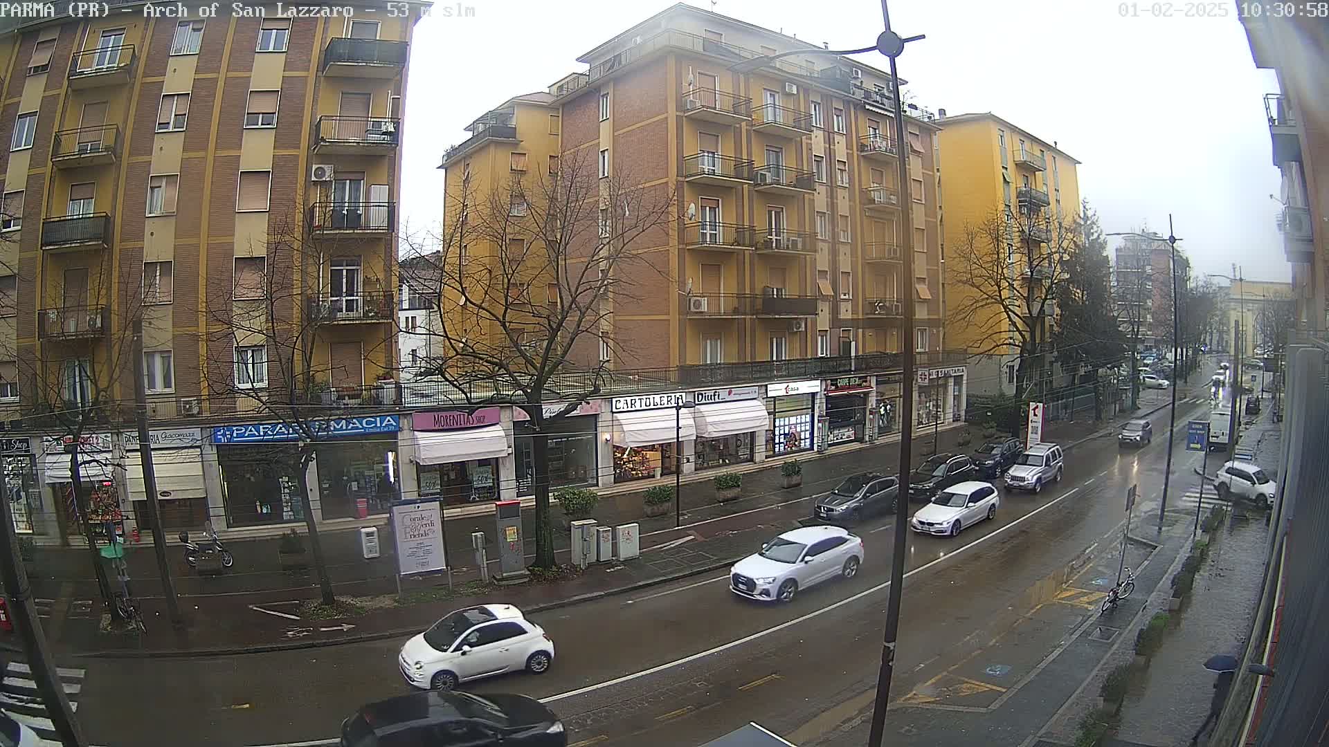 A rain-slicked city street shows a multi-story yellow building with shops on the ground floor, several cars driving, and pedestrians on the sidewalk.