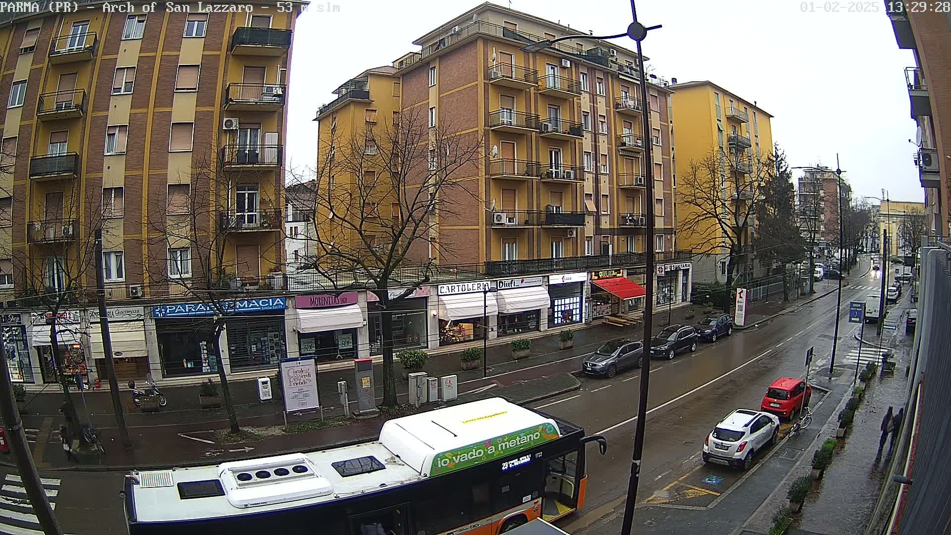 A wet city street scene shows a row of multi-story buildings with shops on the ground floor, several parked cars, a bus, and a few pedestrians under an overcast sky.