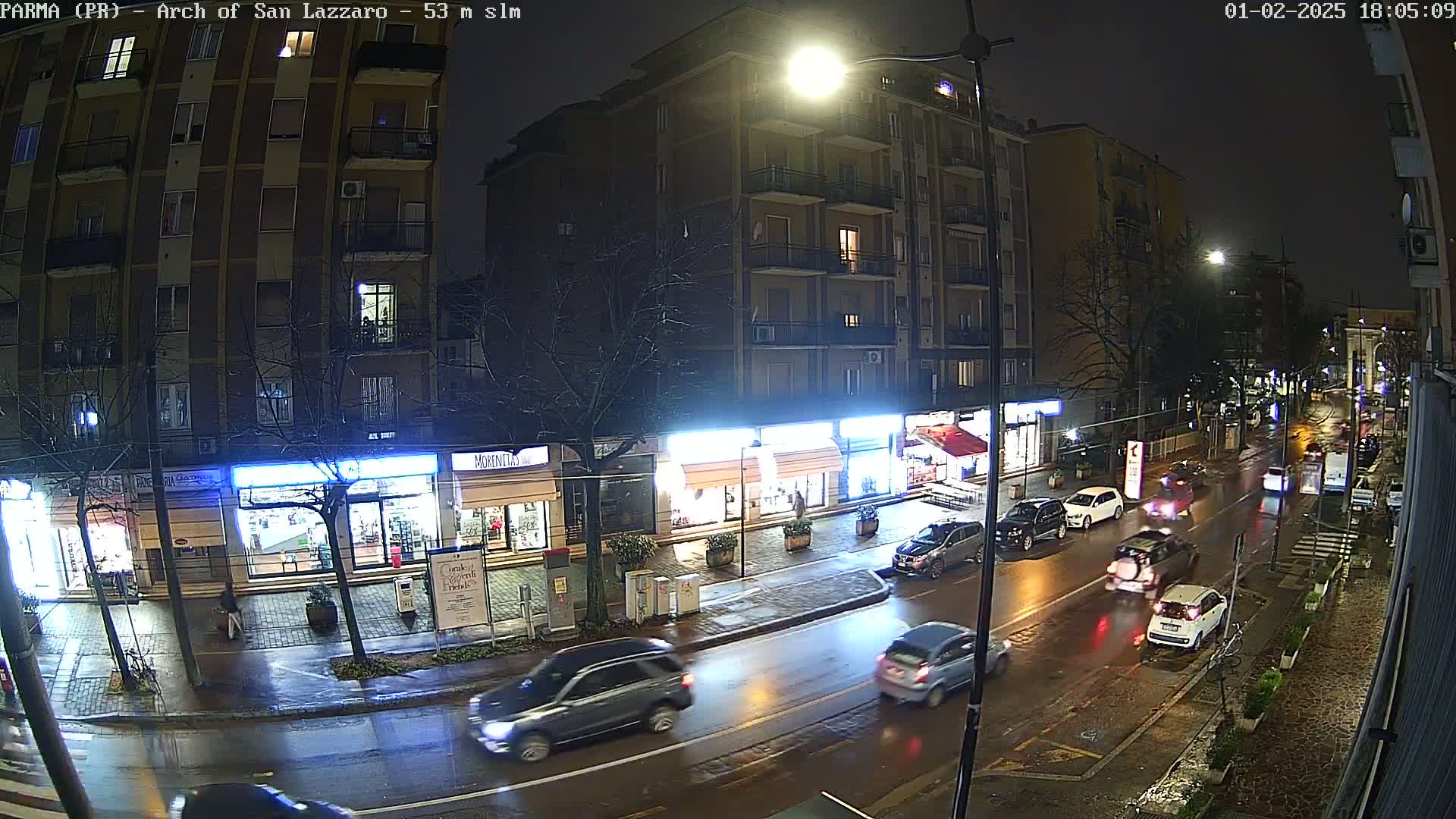 A rain-slicked city street at night, with cars driving past shops and buildings under bright streetlights.