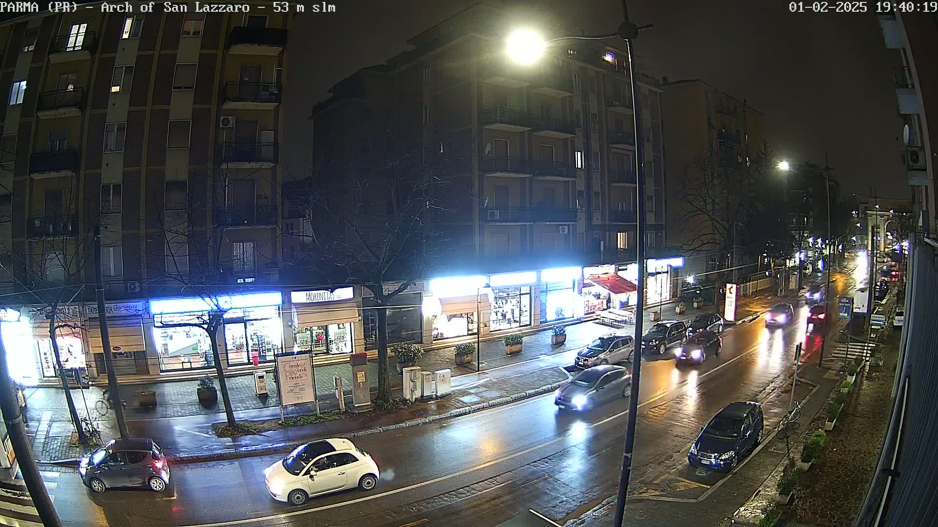 A wet, nighttime street scene shows several cars driving past shops and apartment buildings under bright streetlights.