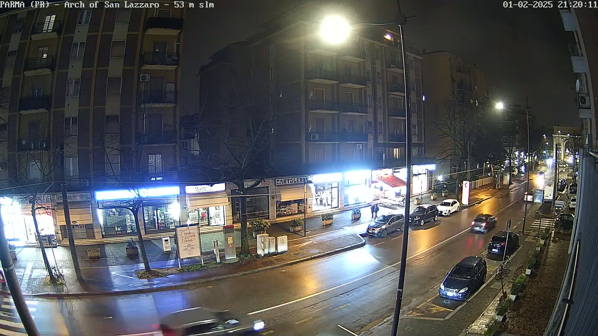 A wet, nighttime street scene shows several cars driving past shops and a multi-story building under bright streetlights.