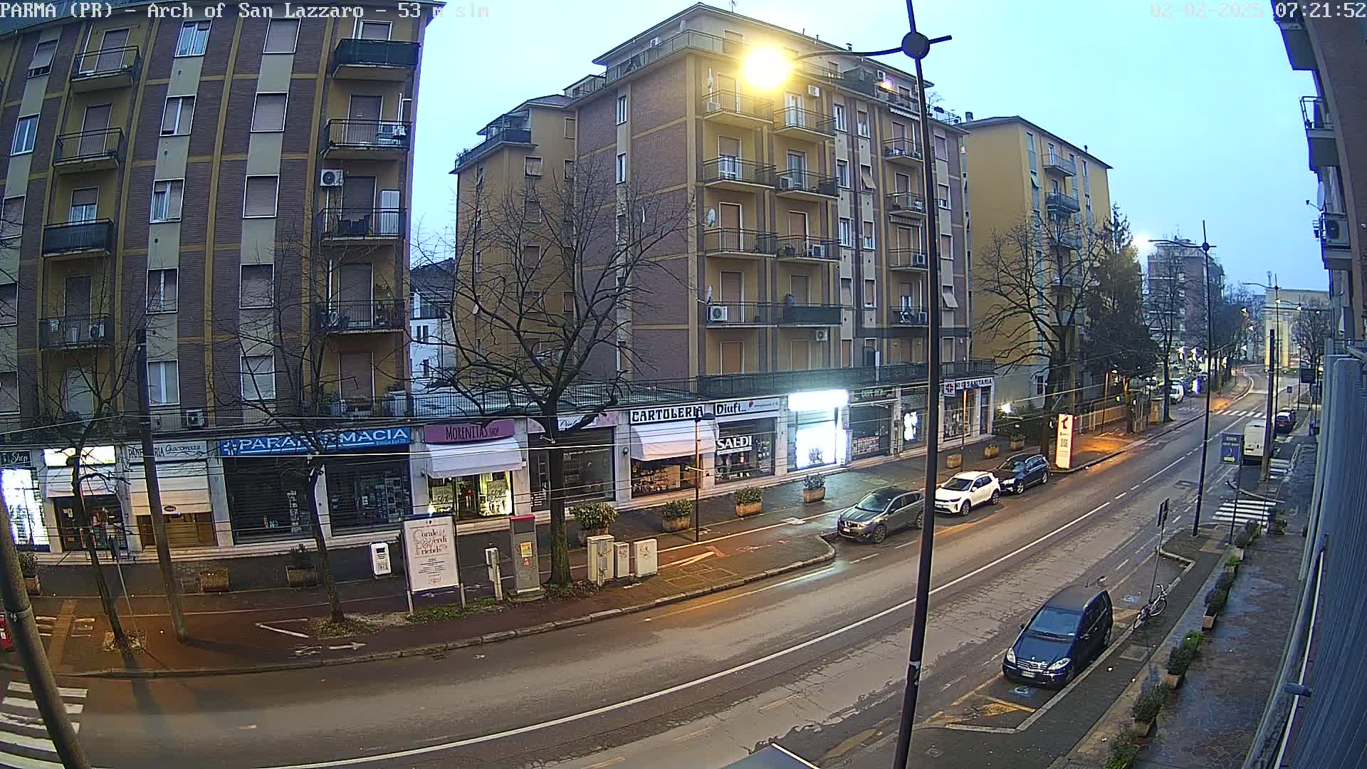 A street scene at dusk shows a row of shops and apartment buildings under a clear, twilight sky, with several cars parked along the side of the road.