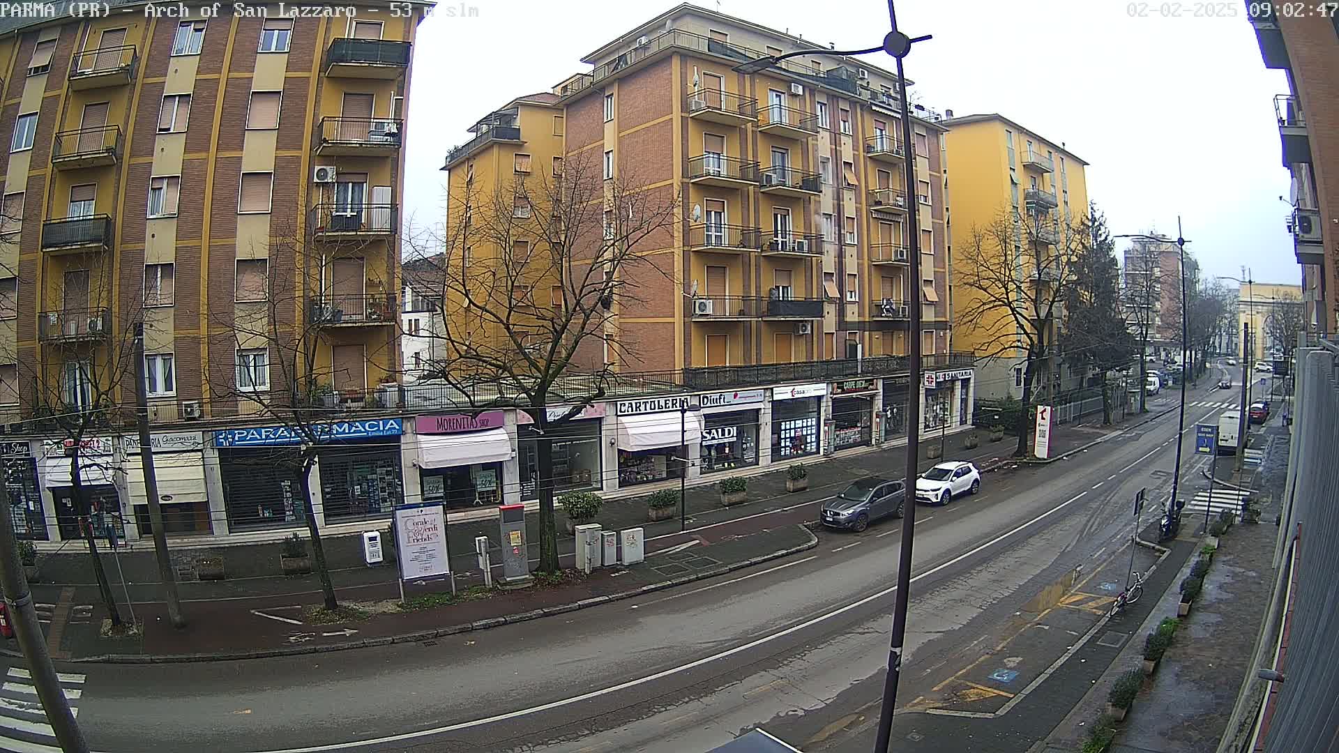 A street scene on an overcast day shows a row of shops and a tall yellow building, with cars parked along the side of the road and a few cars driving down the street.