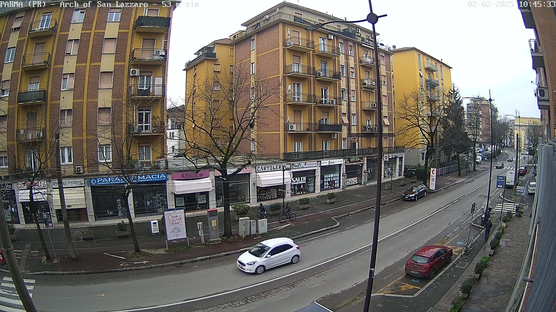A city street scene on an overcast day shows a multi-story yellow building and several shops along a road with cars and a few pedestrians.