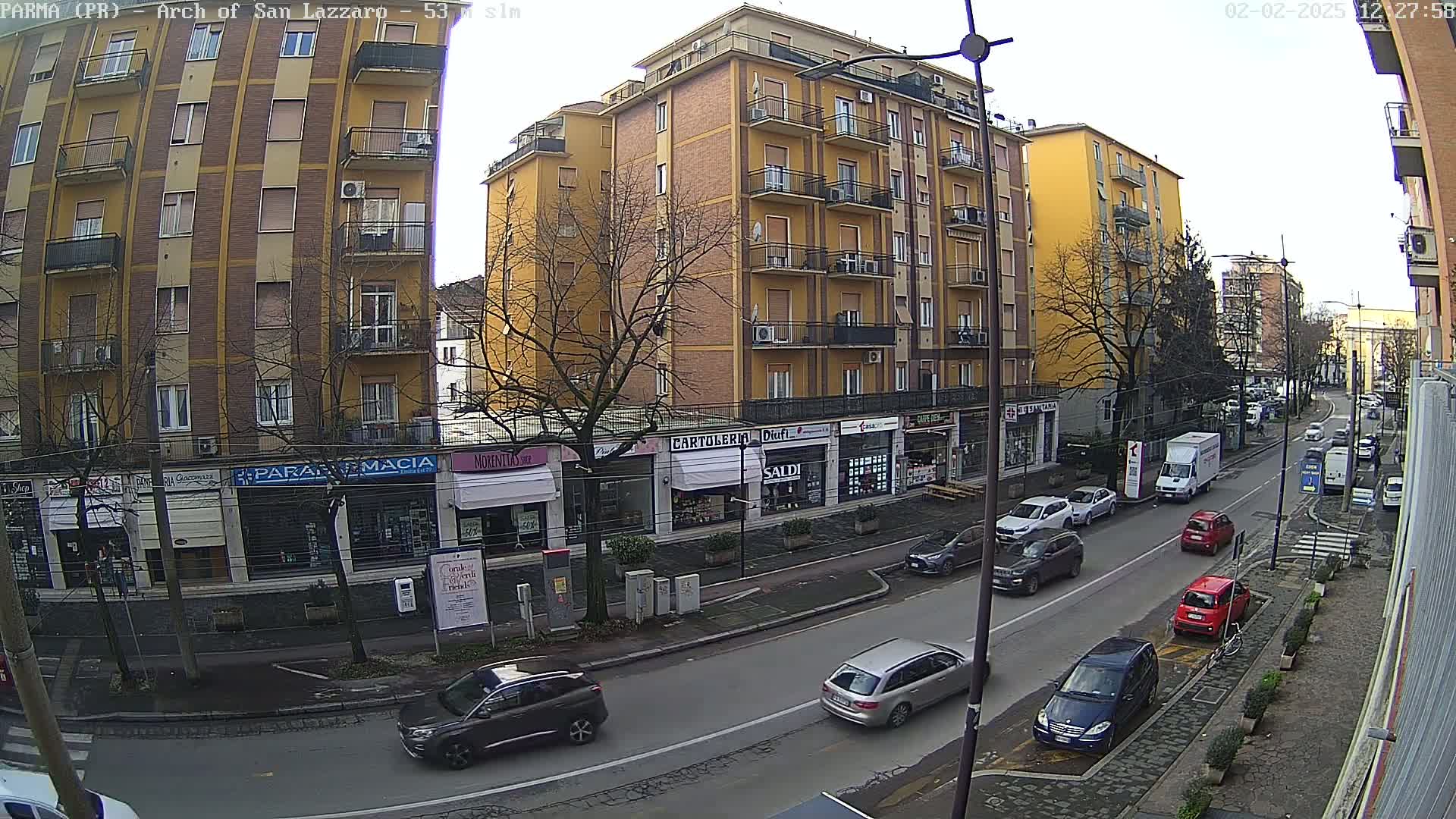 A city street scene on an overcast day shows several cars parked and driving along the street in front of multi-story buildings with shops on the ground floor.