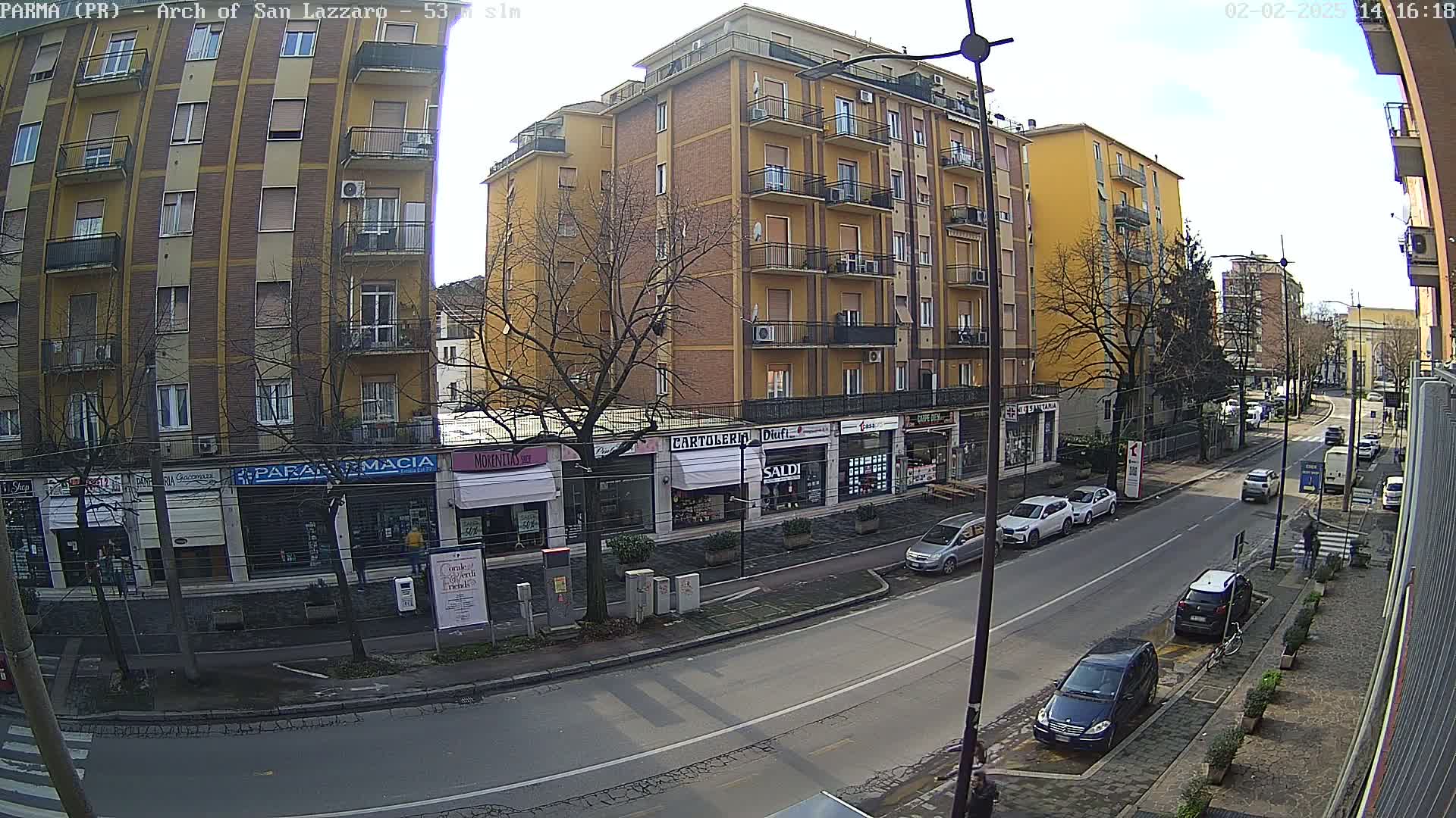 A sunny city street scene shows several multi-story buildings, shops, parked cars, and a few cars driving down the road.