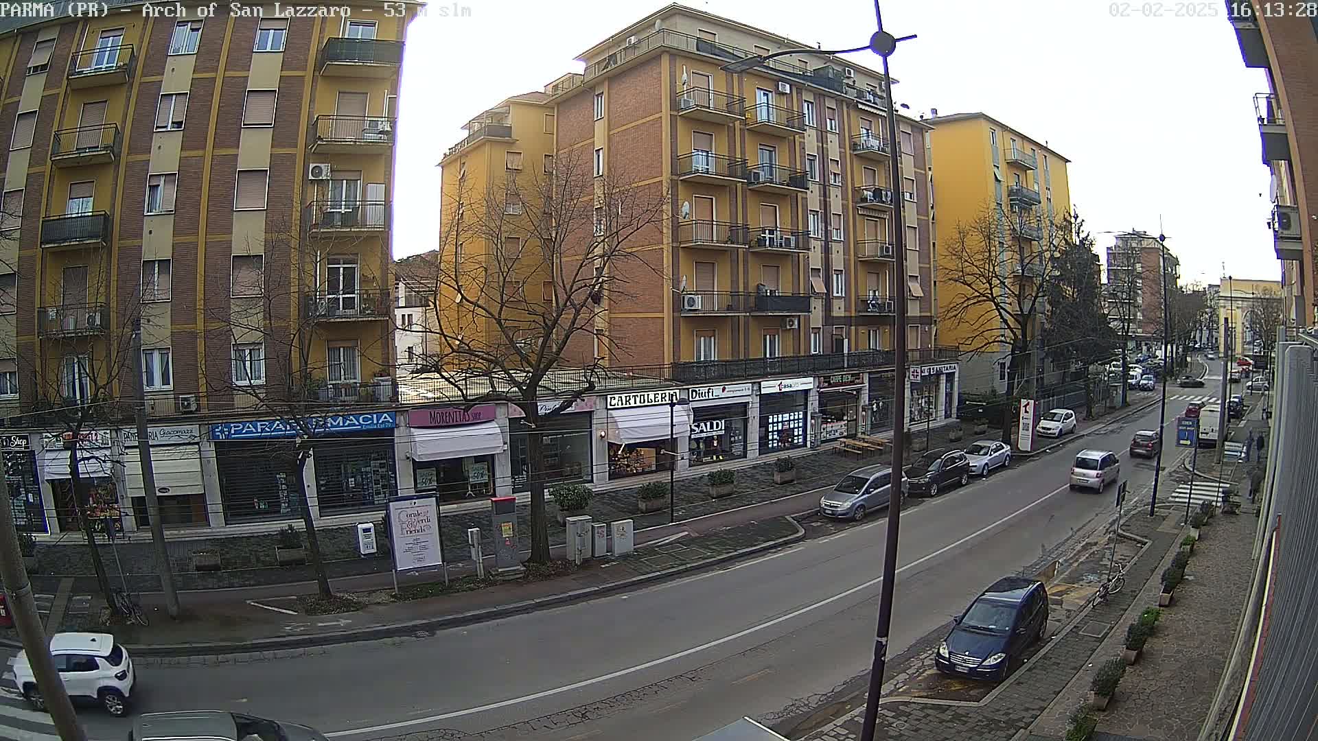 A street scene on an overcast day shows several multi-story buildings and parked cars along a road with sparse traffic.