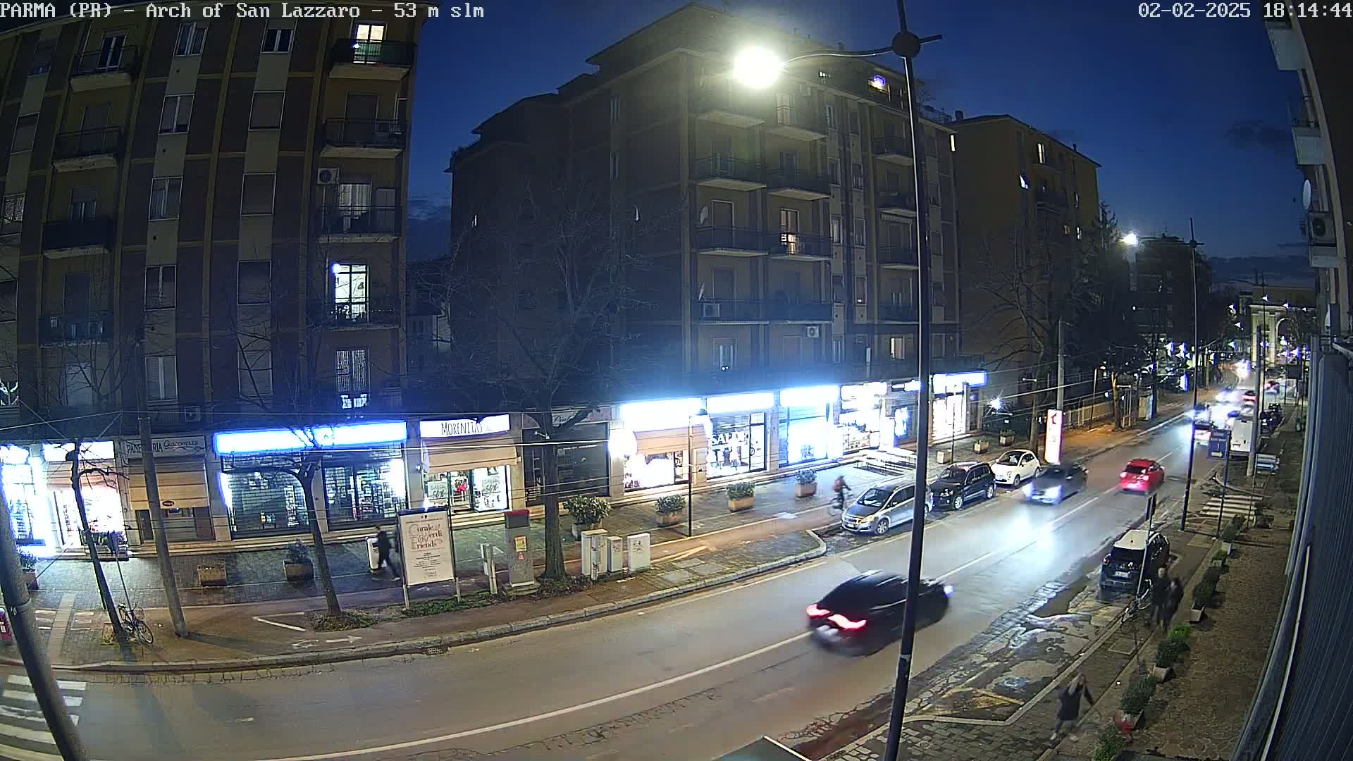 A nighttime street scene shows several cars driving on a roadway lined with multi-story buildings and storefronts under a dark blue, twilight sky.