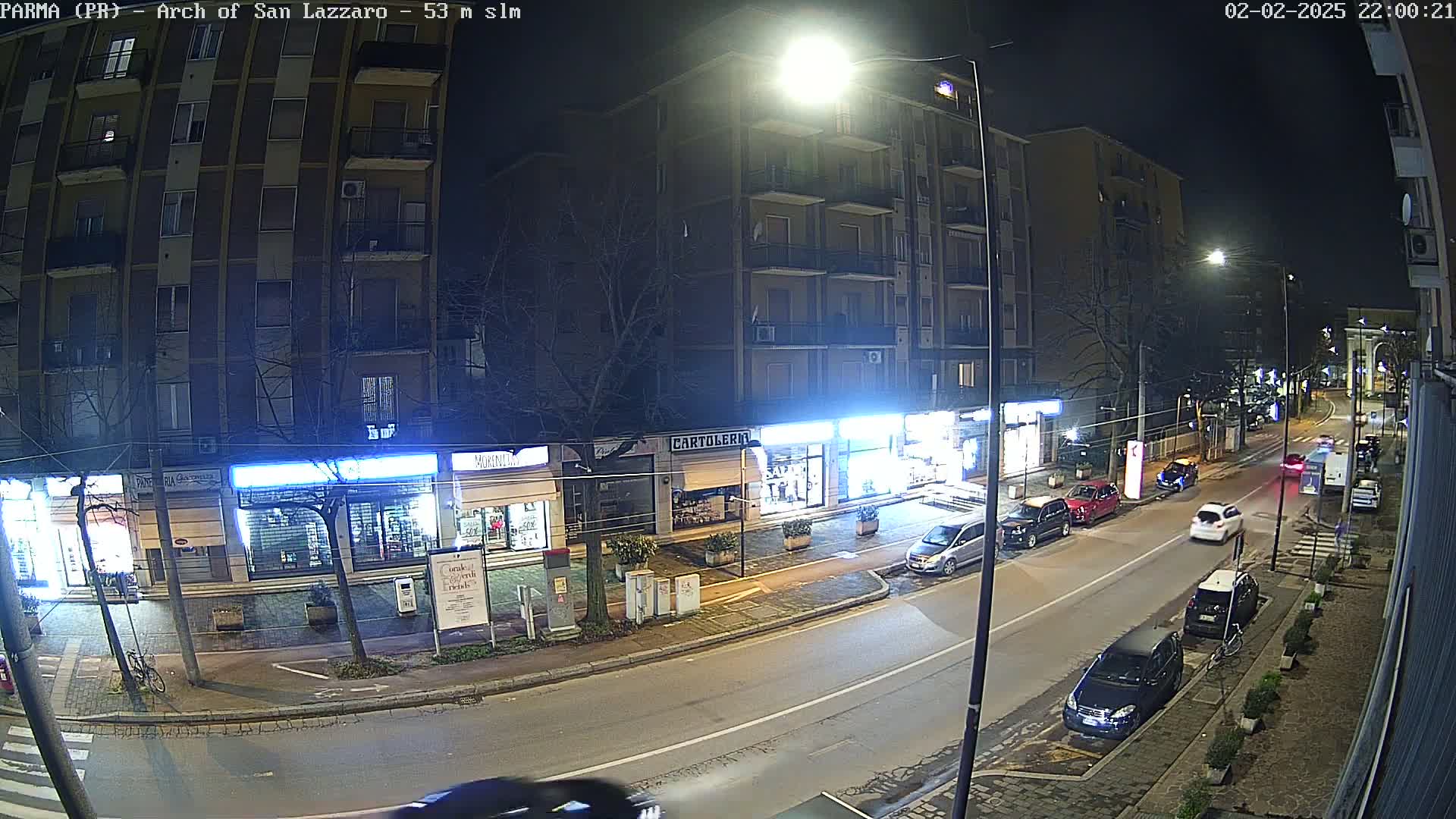A nighttime street scene shows several parked and moving cars along a road lined with multi-story buildings and shops under bright streetlights.
