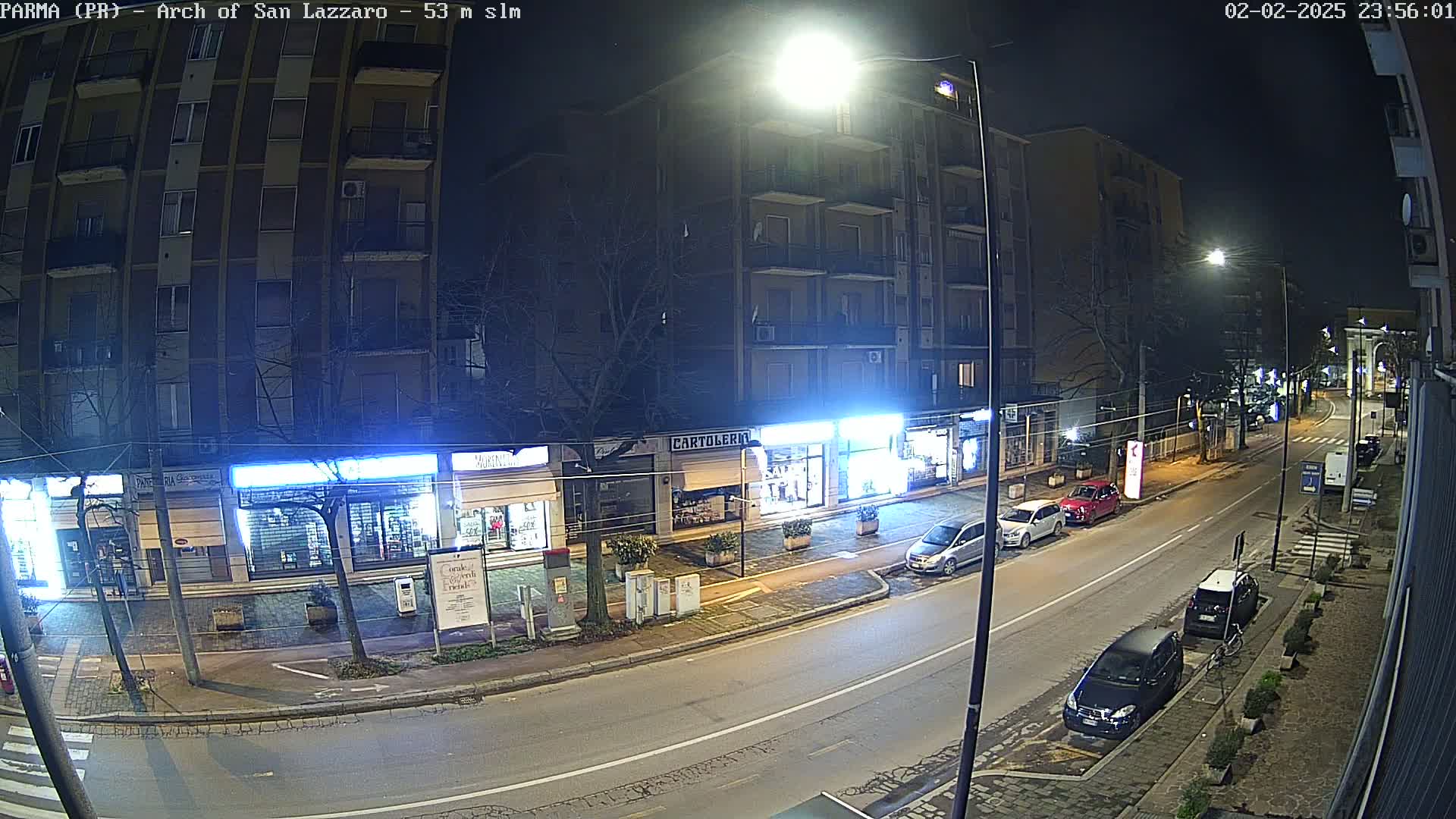A nighttime street scene shows a multi-story building and several parked cars along a roadway under bright streetlights.