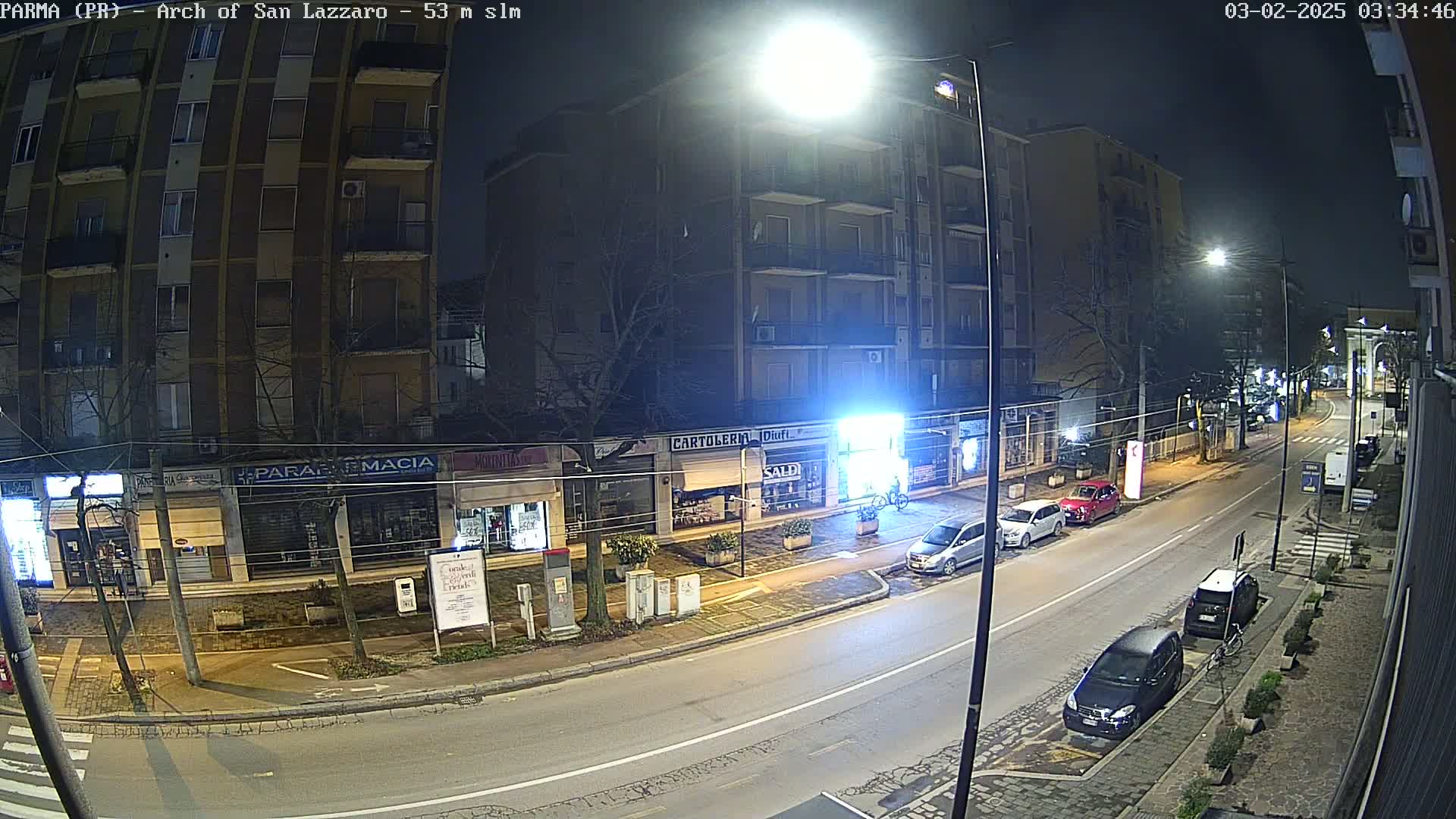 A nighttime street scene shows several parked cars along a roadway lined with shops and apartment buildings under bright streetlights.