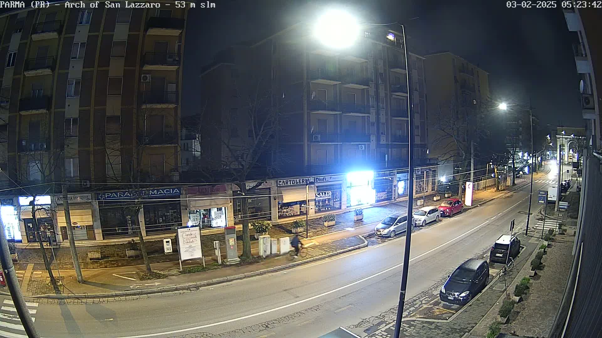 A nighttime street scene shows a multi-story building and several shops with parked cars along the road under bright streetlights.