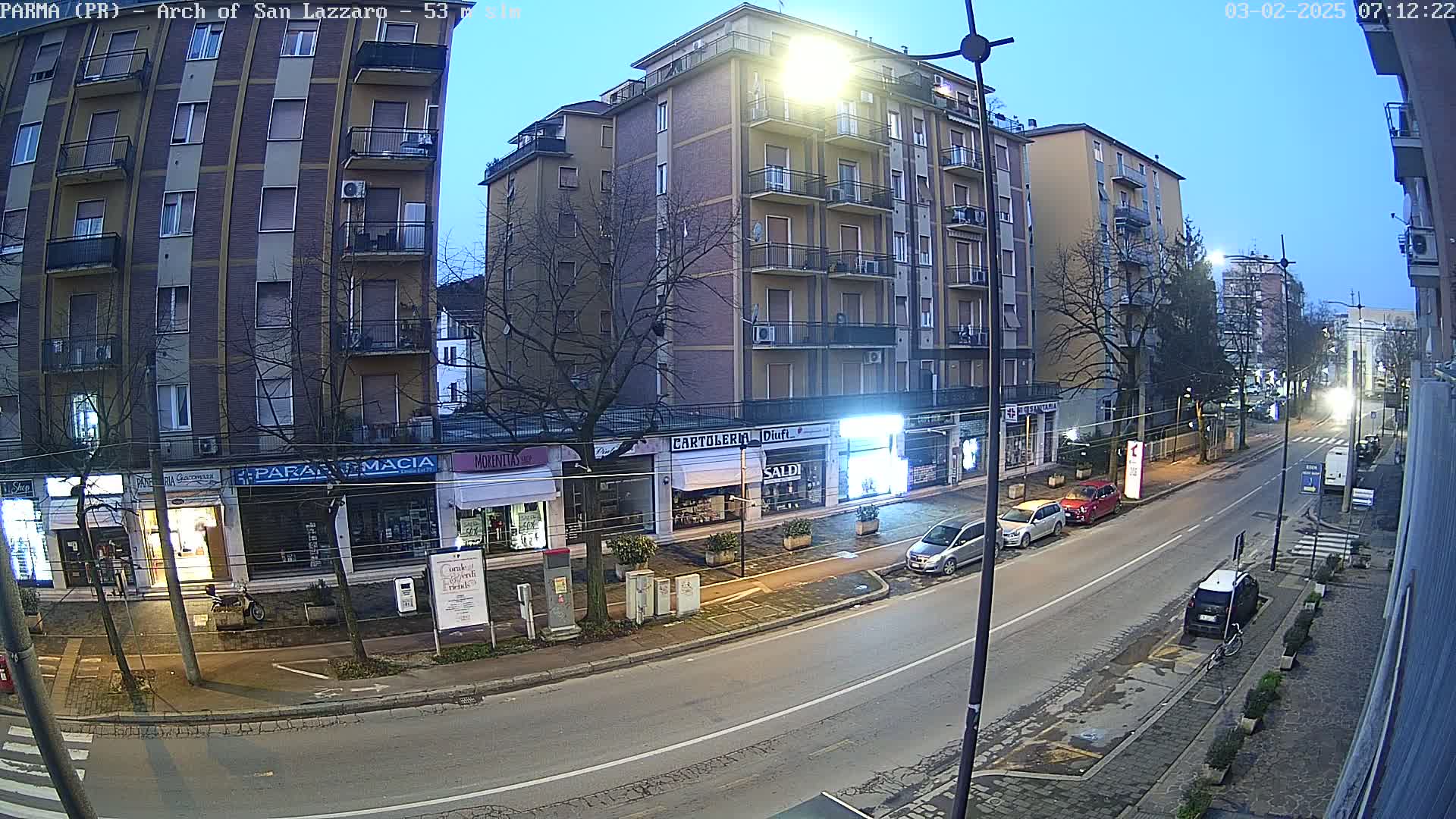 A street scene at twilight shows a row of shops and apartment buildings lining a relatively quiet road with several parked cars.