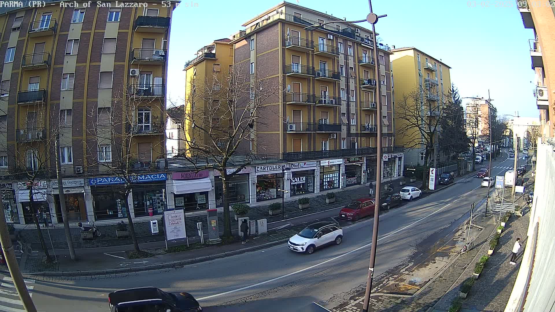 A sunny day on a city street shows several multi-story buildings, shops at street level, parked and moving cars, and bare winter trees.