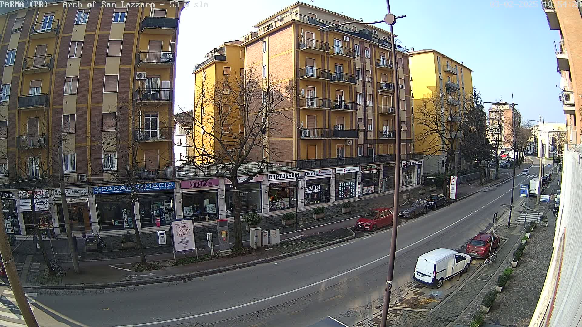A sunny city street scene shows a multi-story yellow building with storefronts on the ground floor, parked cars lining the street, and a few pedestrians.