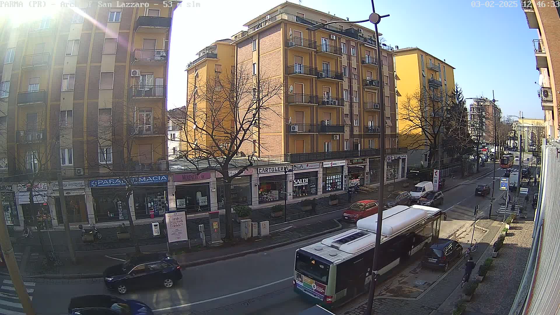A sunny day on a city street shows a yellow multi-story building with shops on the ground floor, several cars, and a bus.