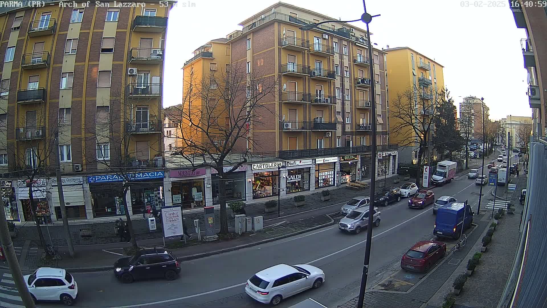A street scene on a partly cloudy day shows a multi-story yellow building with shops on the ground floor, several cars driving on a road, and parked cars along the sides.