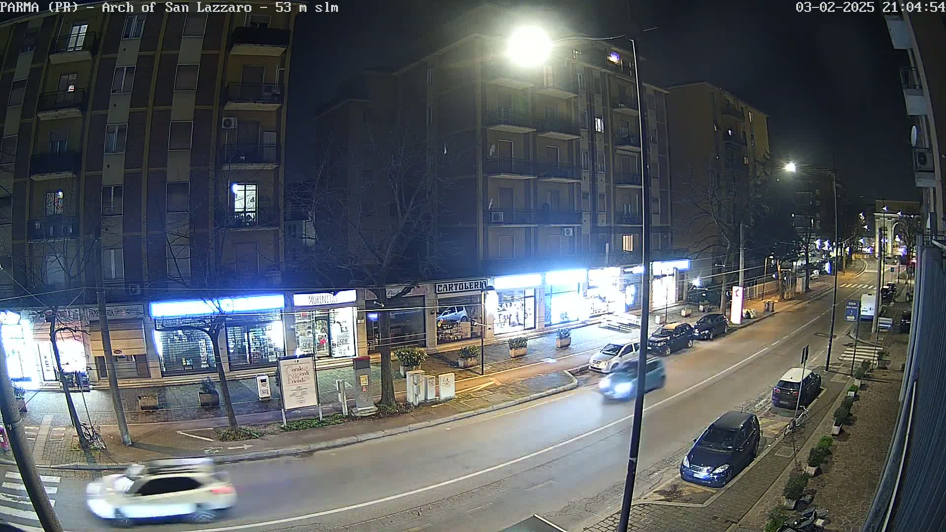 A nighttime street scene shows several cars driving on a road lined with shops and buildings under streetlights.