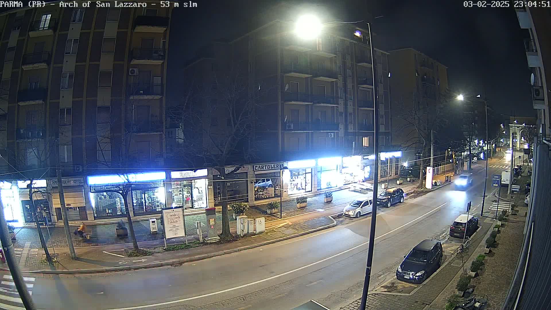 A nighttime street scene shows a multi-story building with shops on the ground floor, several parked cars, and a few cars driving on a two-lane road.