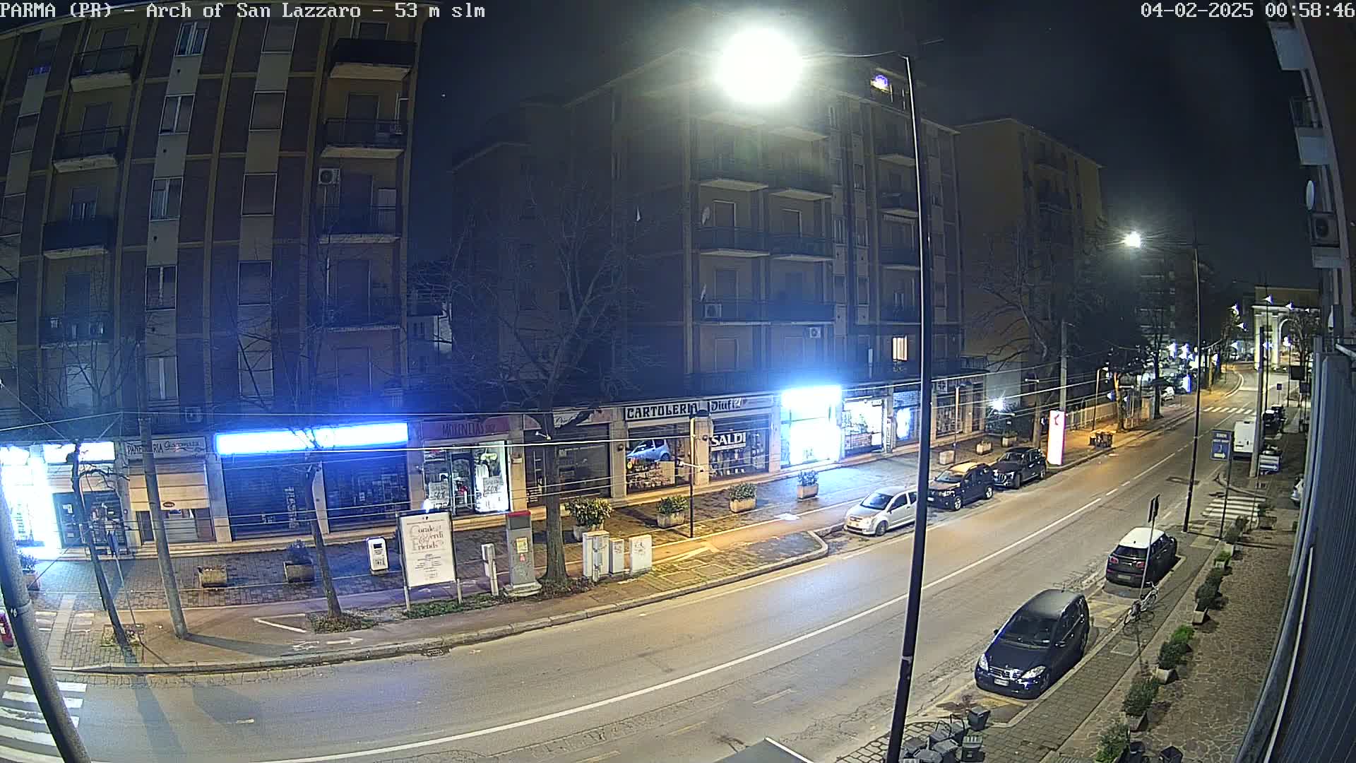A nighttime street scene shows a multi-story building with storefronts and parked cars along a roadway under bright streetlights.