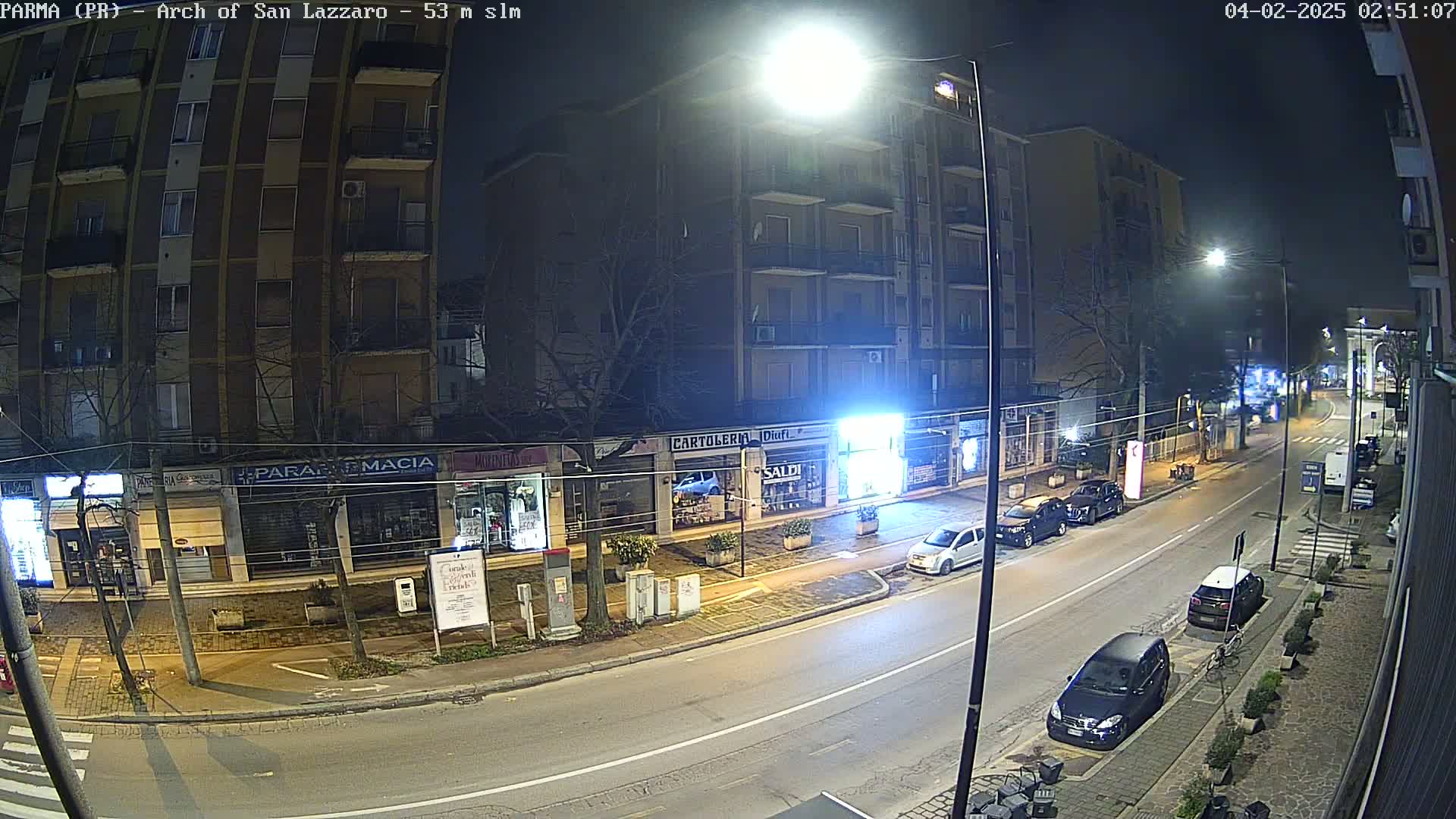 A nighttime street scene shows a multi-story building with storefronts on the ground floor, several cars parked along the curb, and a mostly empty road.