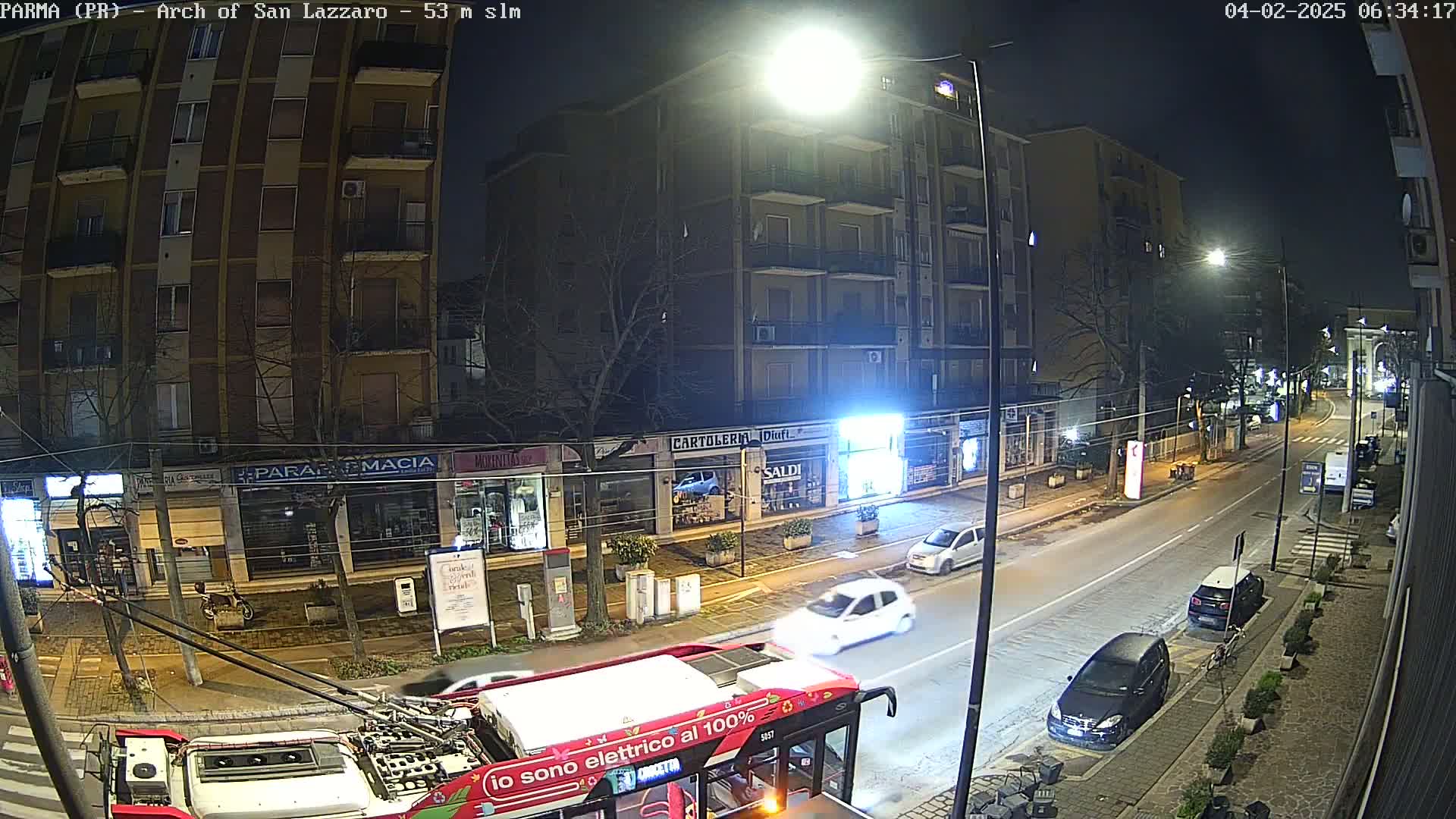 A nighttime street scene shows a multi-story building and several parked and moving cars, viewed from the perspective of a nearly-frontal bus.