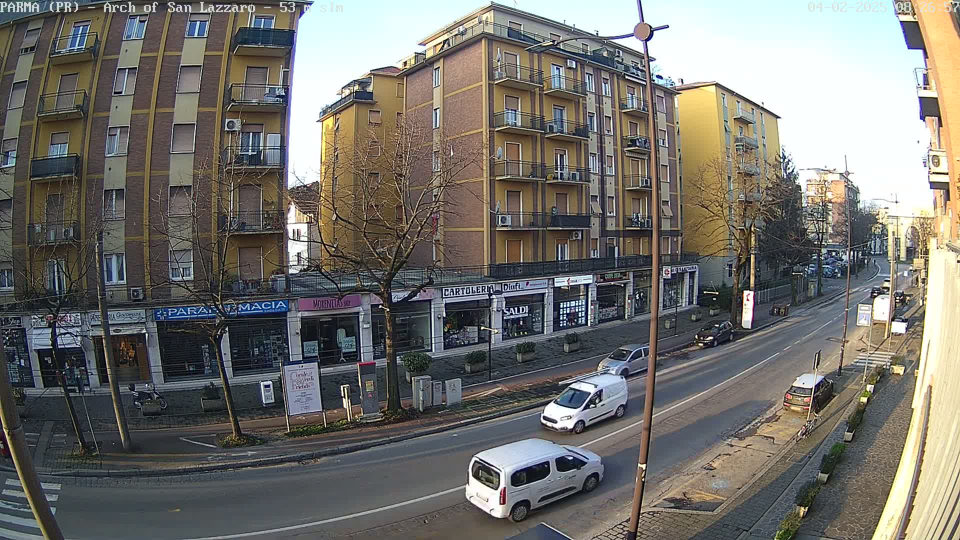 A sunny day reveals a street scene in a European city, where several cars are parked and driving along a road lined with multi-story buildings and shops.