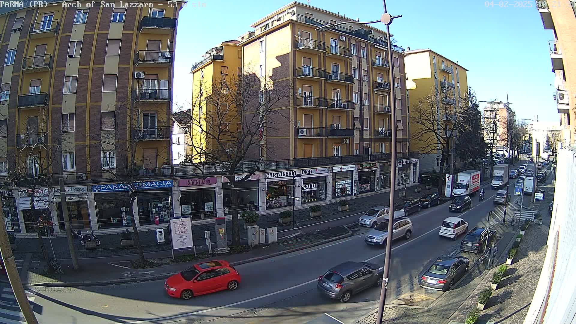 A sunny city street scene shows a multi-story yellow building and several shops along a roadway with moderate traffic.