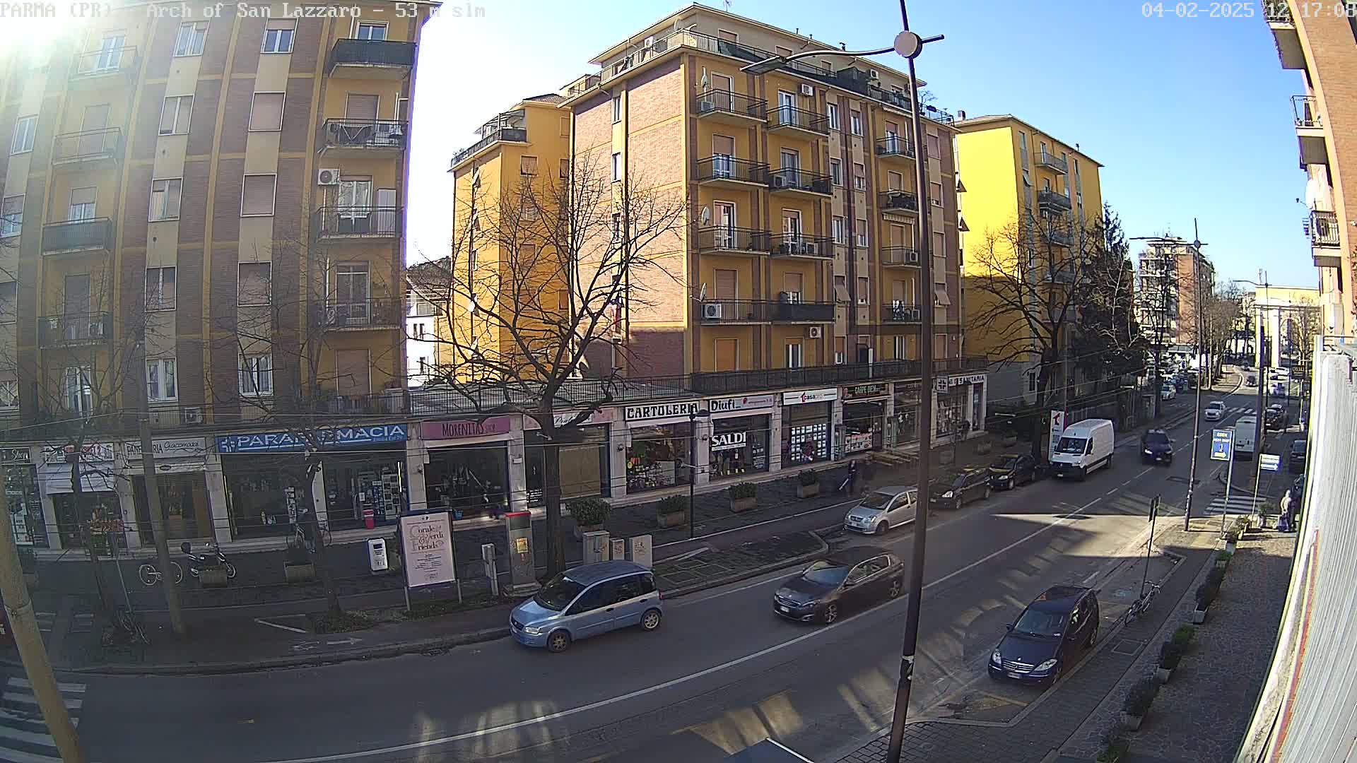 A sunny day on a city street with cars parked and driving past several multi-story buildings and shops.