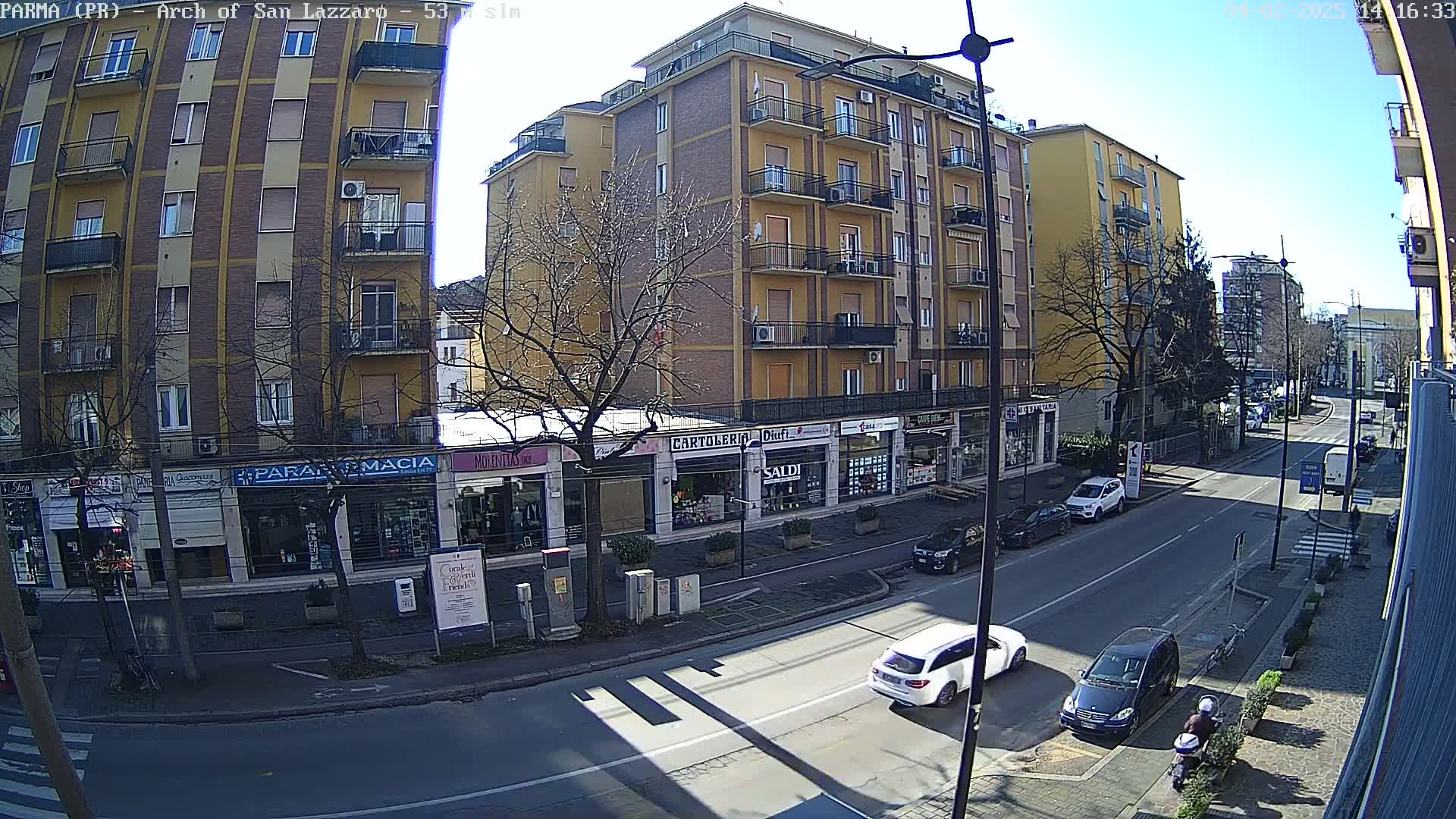 A sunny day reveals a street scene with several cars parked and driving along a multi-lane road lined with multi-story buildings containing various shops.