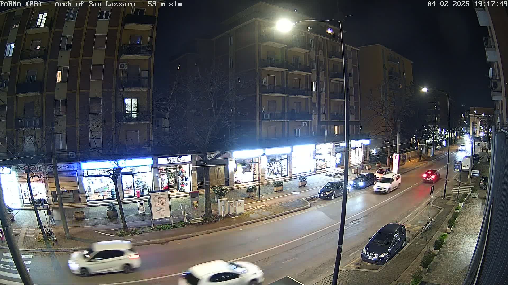 A nighttime street scene shows several cars driving on a road lined with shops and a multi-story building under streetlights.