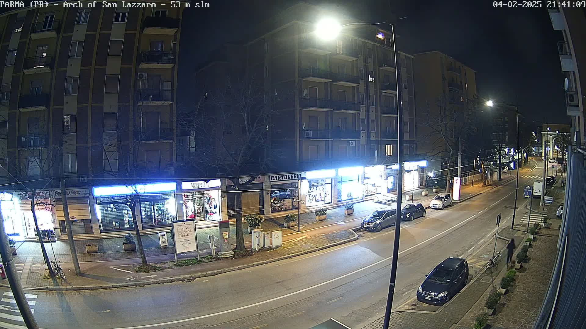 A nighttime street scene shows a multi-story building with storefronts, cars parked along the side of the road, and several people walking.