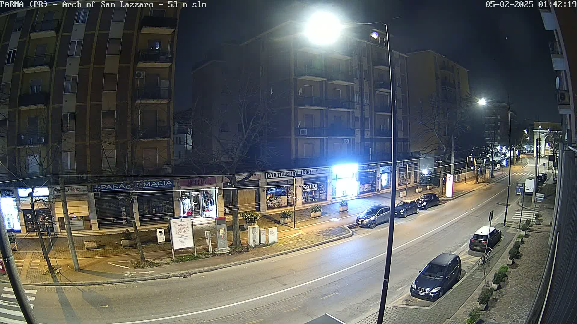 A nighttime street scene shows several parked cars and a few buildings with illuminated storefronts.