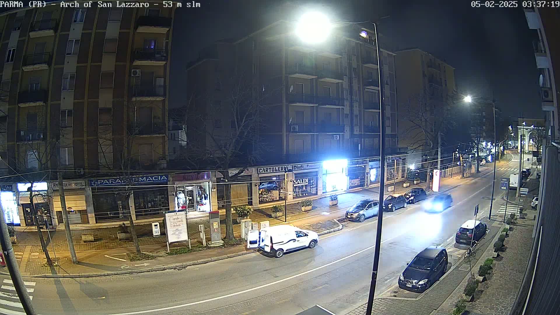 A nighttime street scene shows several cars parked and driving along a road lined with multi-story buildings and shops.