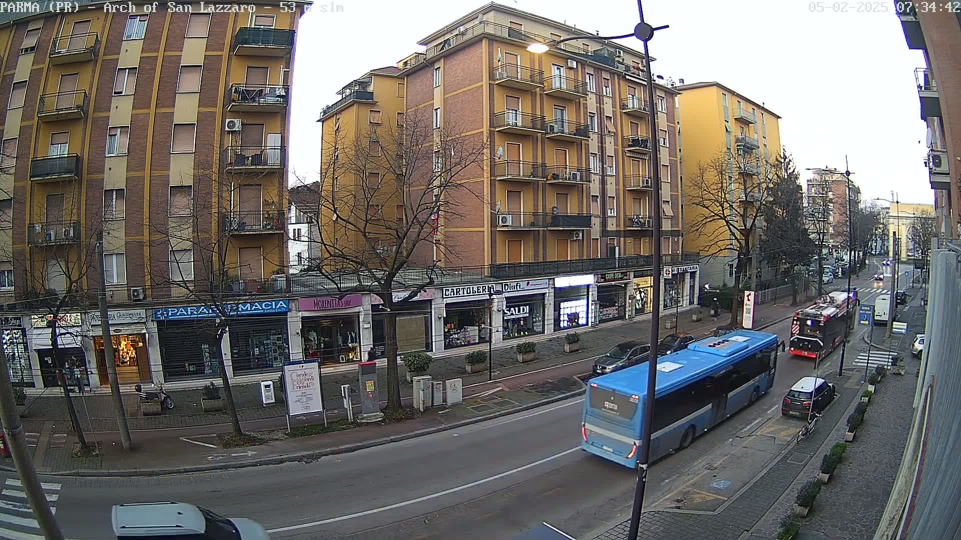 A blue bus drives down a city street lined with multi-story buildings under an overcast sky.