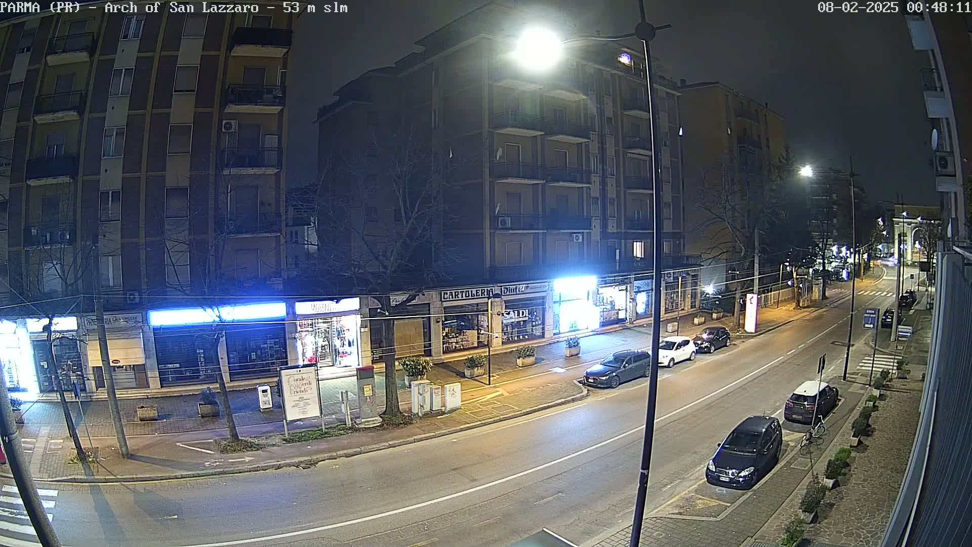 A nighttime street scene shows several parked cars along the curb in front of shops and apartment buildings under bright streetlights.