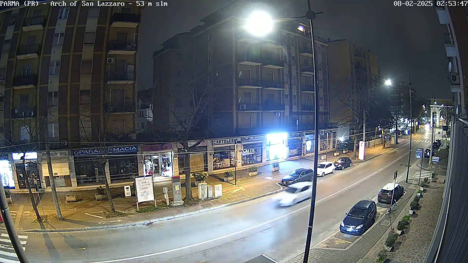 A nighttime street scene shows several cars parked and driving past multi-story buildings with shops on the ground floor under bright streetlights.