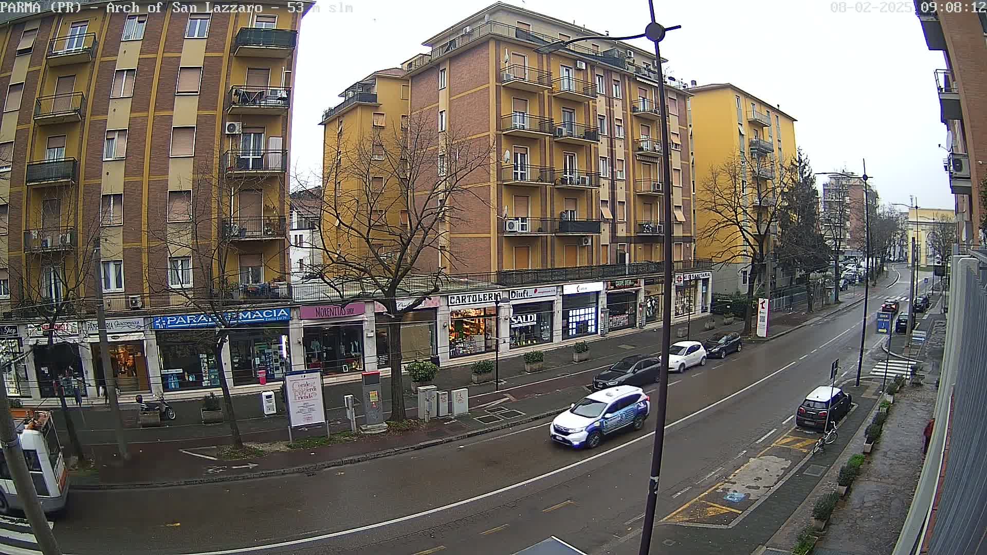 A street scene on an overcast day shows a row of multi-story buildings with shops on the ground floor, several cars parked and driving along the street, and bare trees lining the sidewalks.
