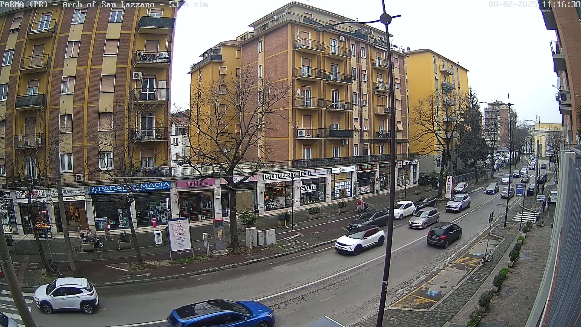 A street scene on an overcast day shows a row of multi-story buildings with shops on the ground floor, and moderate traffic moving along the road in front.