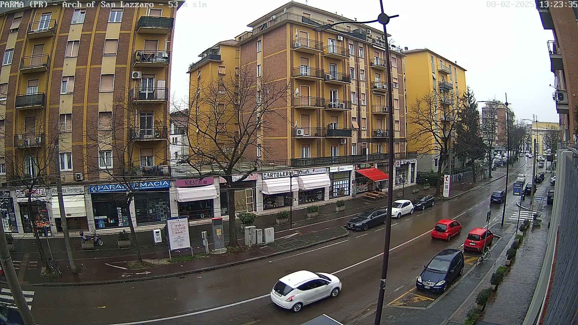 A wet city street scene shows several cars parked and driving past multi-story buildings with shops on the ground floor under an overcast sky.