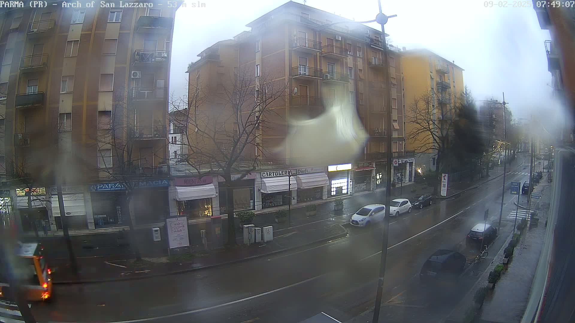 A rain-slicked city street scene shows several multi-story buildings, several cars parked and driving down the street, and some pedestrians.
