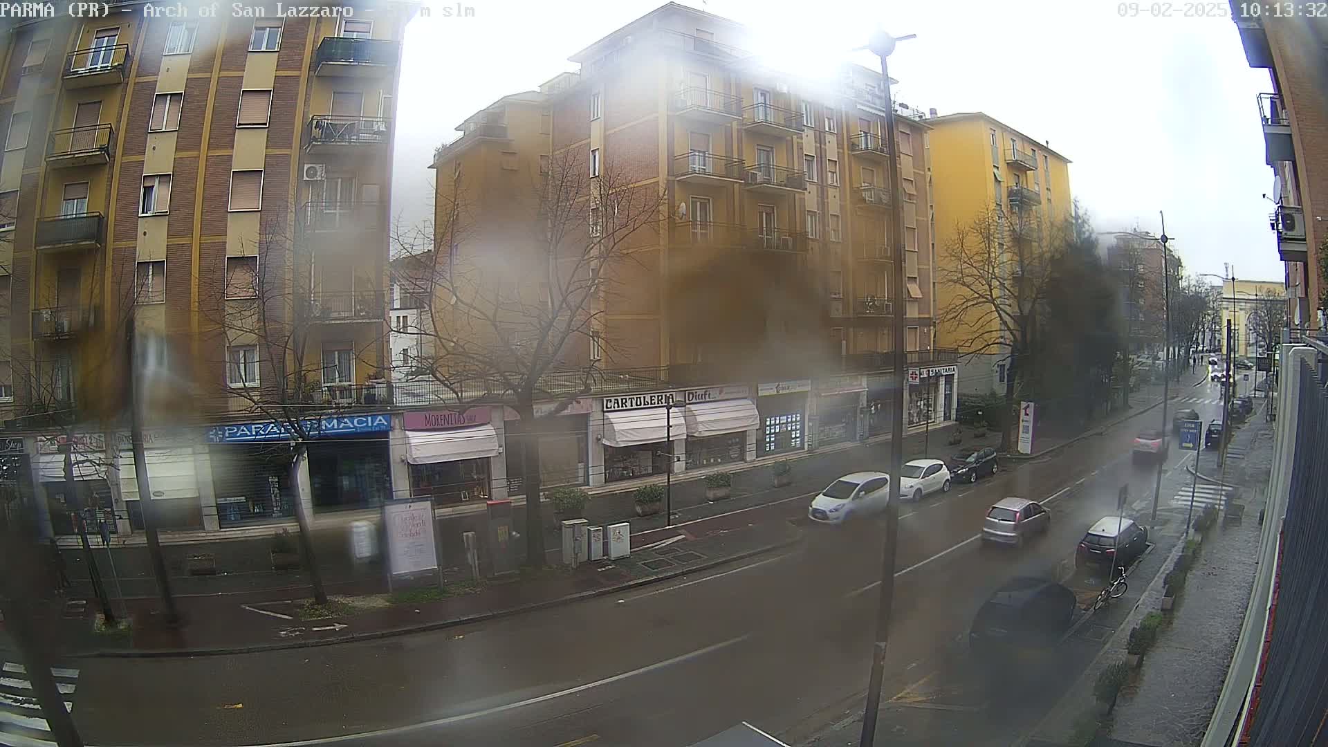 A rain-streaked view from a window shows a city street with several cars and pedestrians, lined with multi-story buildings and shops.