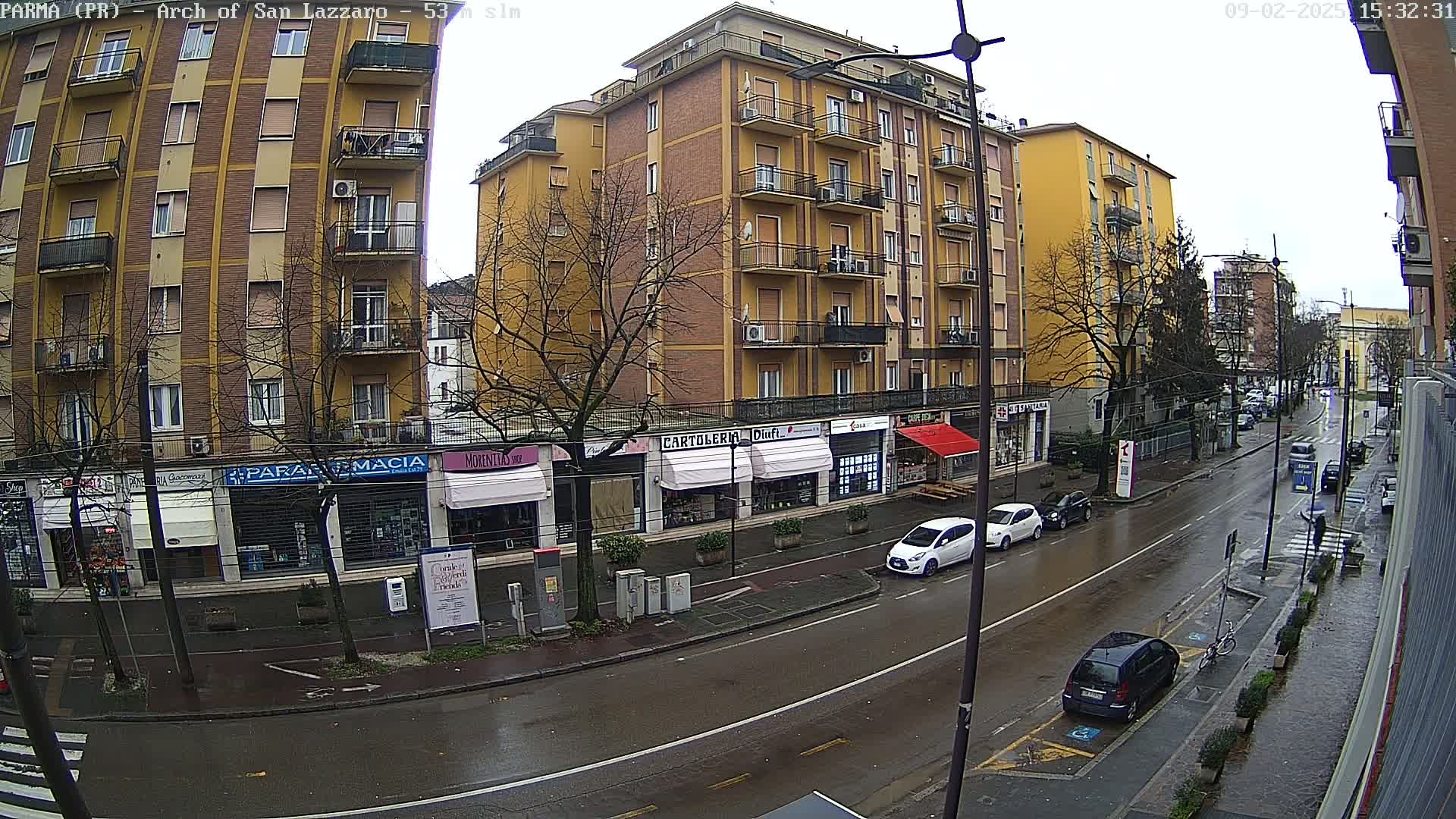 A wet city street scene shows several multi-story buildings with shops on the ground floor, several cars parked and driving along the street, and bare trees in overcast weather.