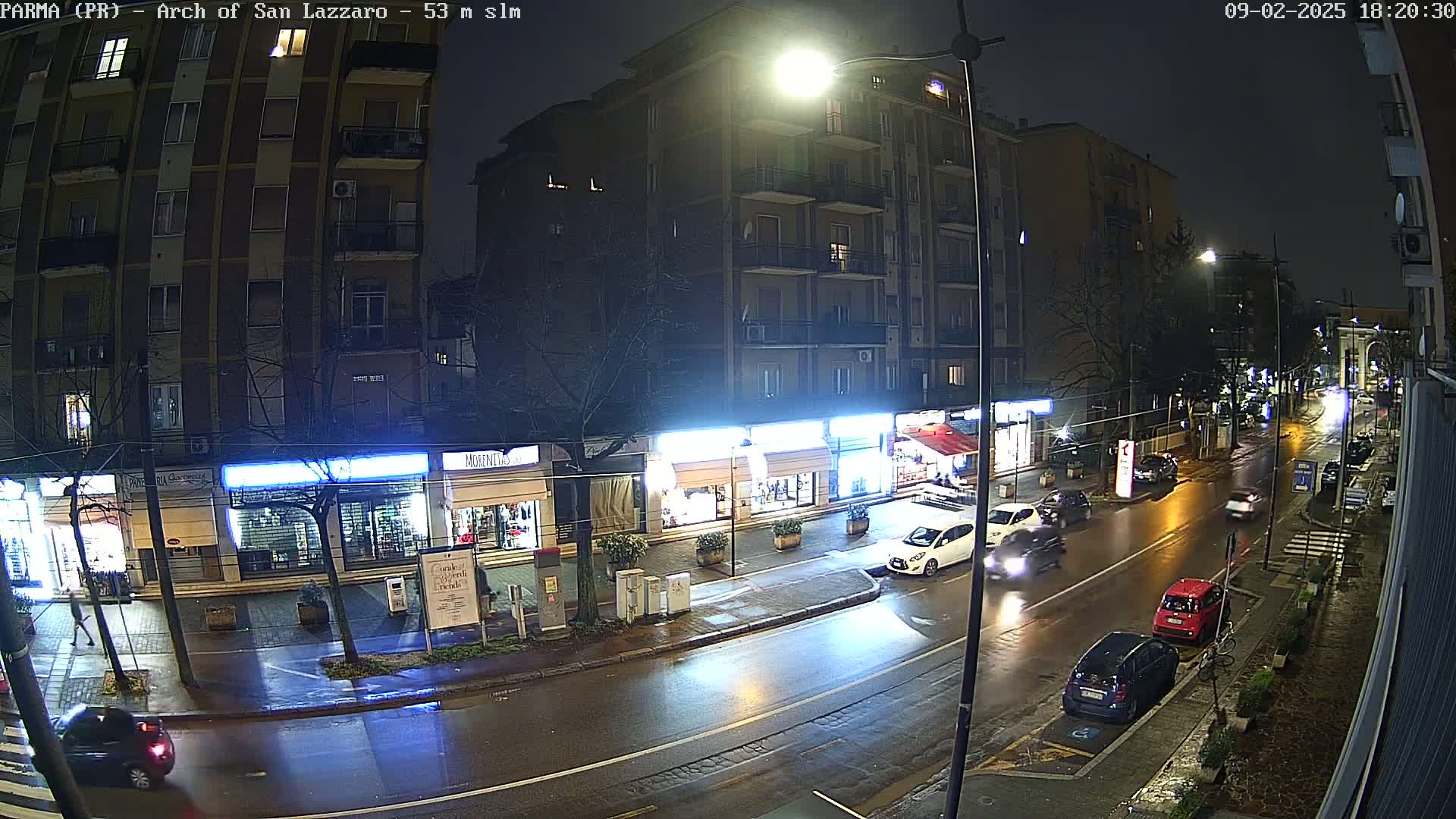 A rain-slicked city street at night, lined with shops and apartment buildings, shows several cars driving along the road.