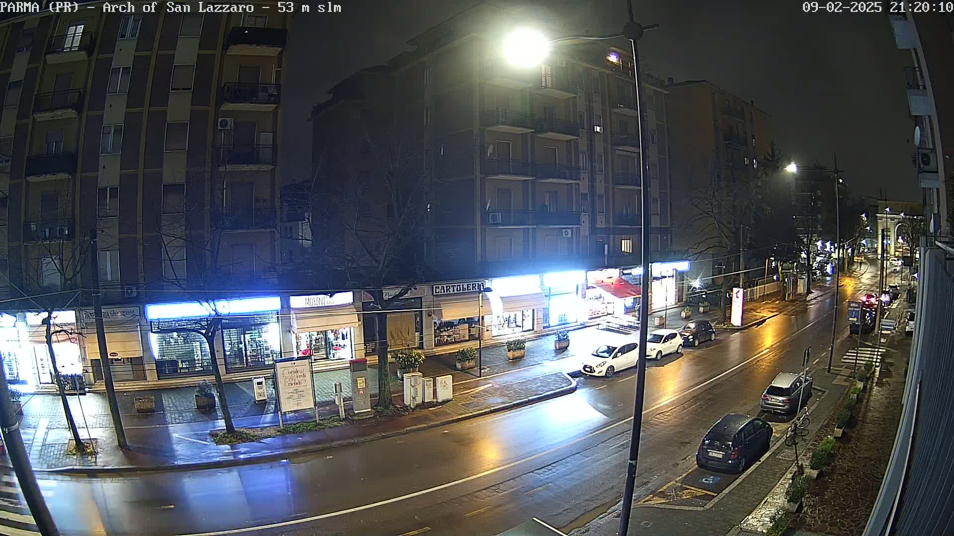 A wet, nighttime street scene shows several parked cars and a few driving cars in front of shops and a multi-story apartment building under bright streetlights.