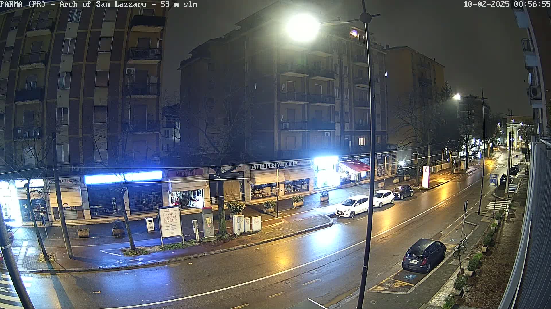 A wet, nighttime street scene shows several cars parked and driving along a road lined with storefronts and apartment buildings under bright streetlights.