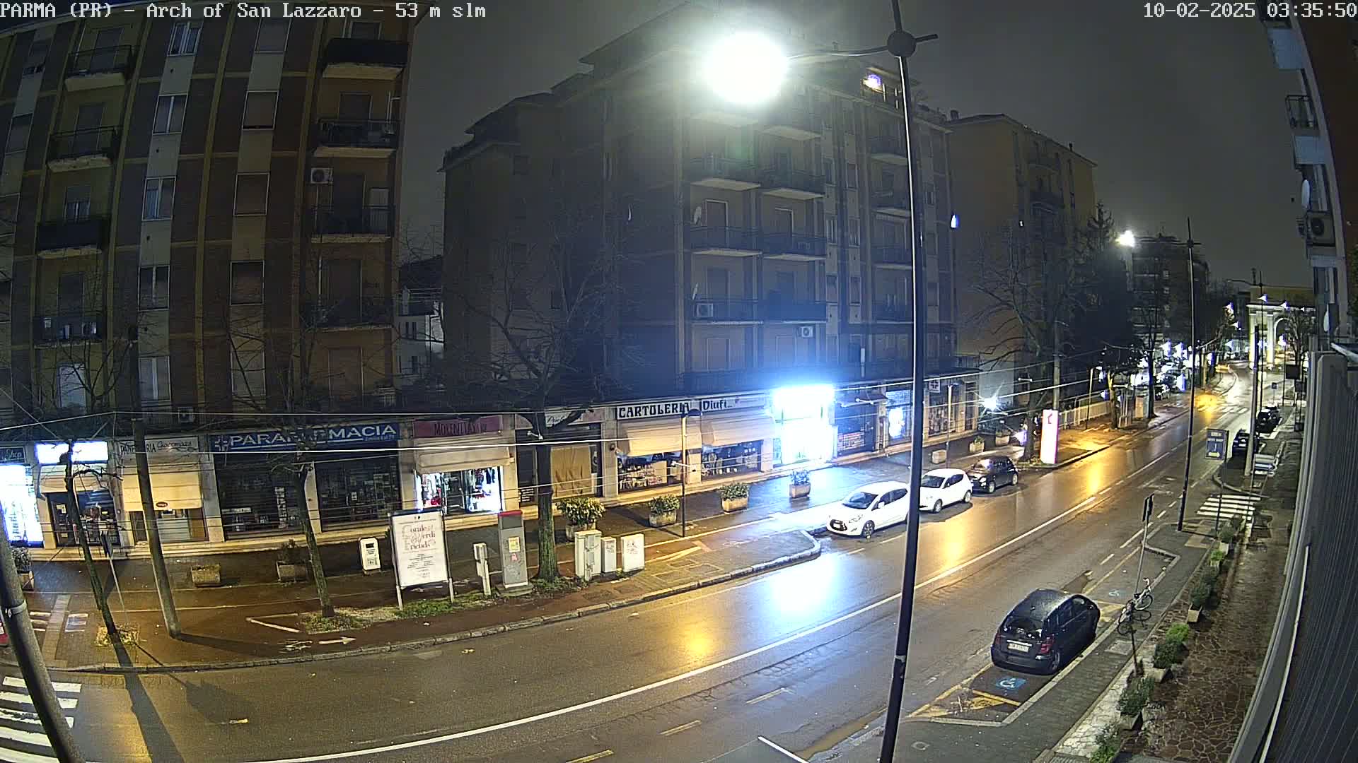 A wet, nighttime street scene shows several cars parked and driving past multi-story buildings and shops.