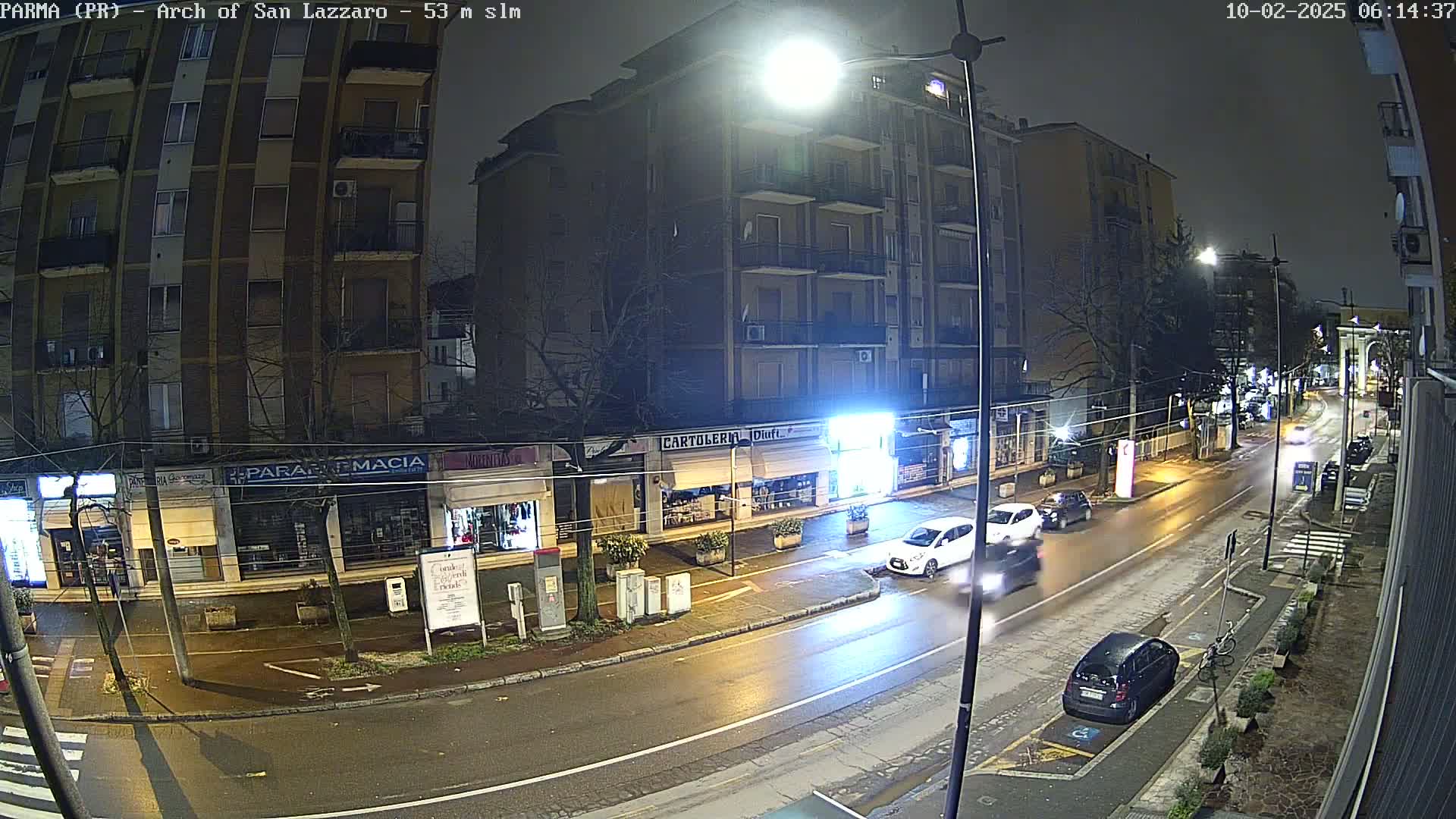 A wet, nighttime street scene shows several cars driving past multi-story buildings with shops on the ground floor, under bright streetlights.