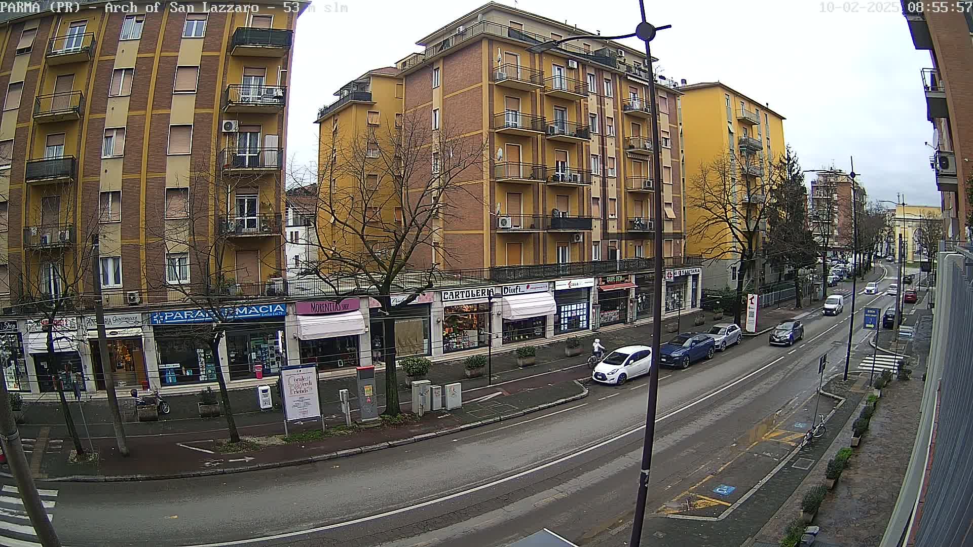 A street scene on an overcast day shows a row of multi-story buildings with shops on the ground floor, and several cars parked and driving along a road.