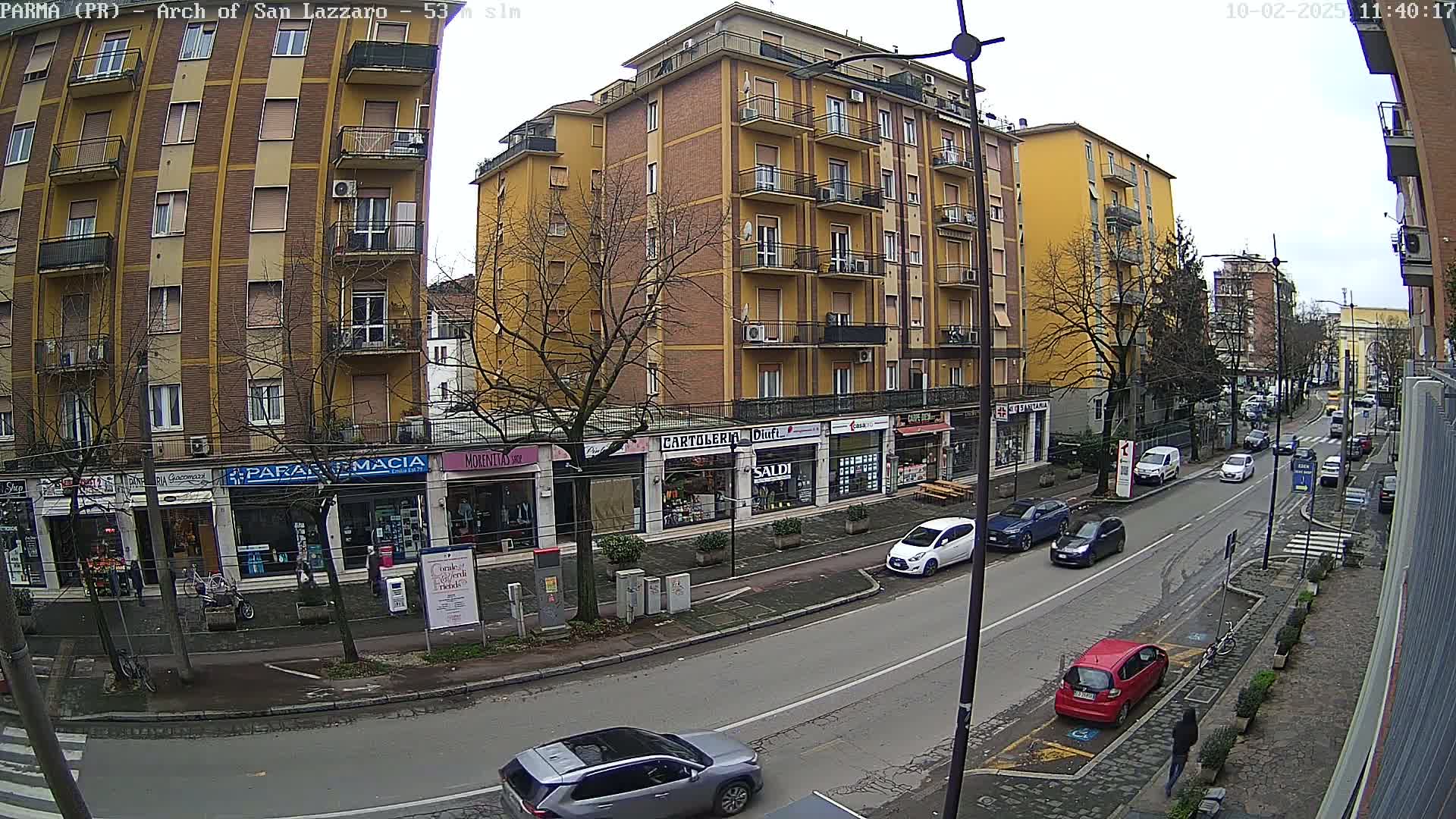 A city street scene on an overcast day shows several multi-story buildings with shops on the ground floor, and various cars parked and driving along the street.